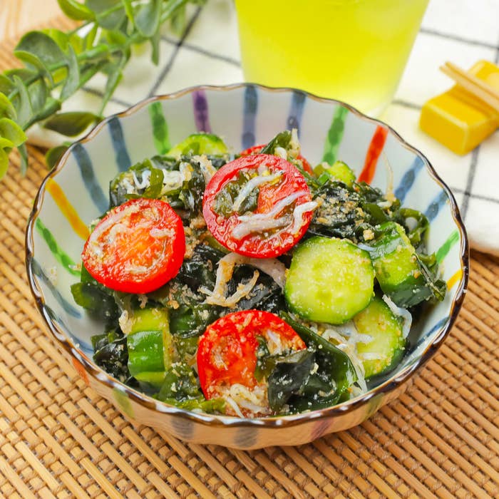 小鉢に盛られた和風サラダ、キュウリ、トマト、ワカメ、ちりめんじゃこが入っている。背景に植物とグラス。