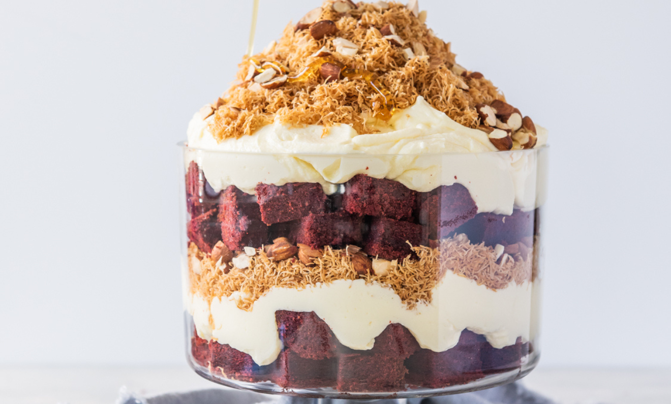 Glass trifle bowl filled with layers of cream, red velvet cake, and a topping of crumbled nuts and shredded pastry