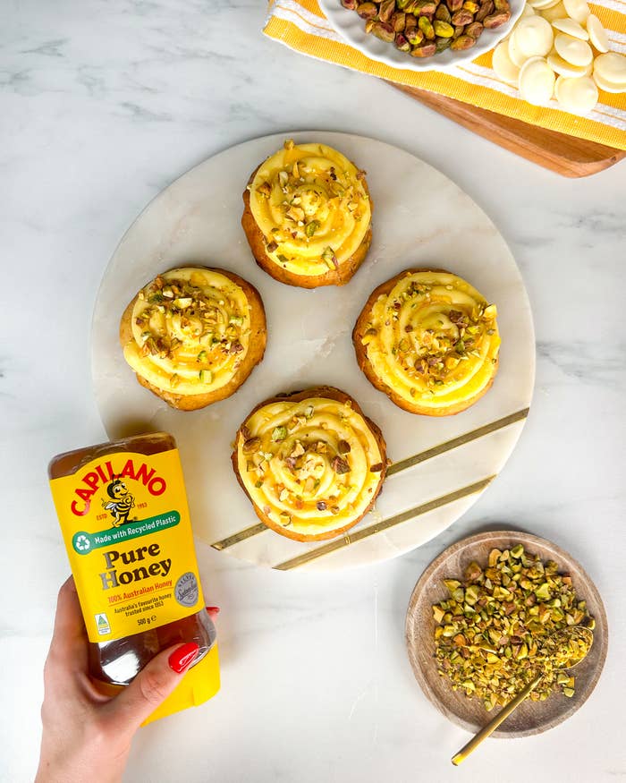 Capilano Pure Honey bottle held above a plate with four desserts topped with pistachios and icing. A dish with white chocolate and a bowl of chopped pistachios are visible