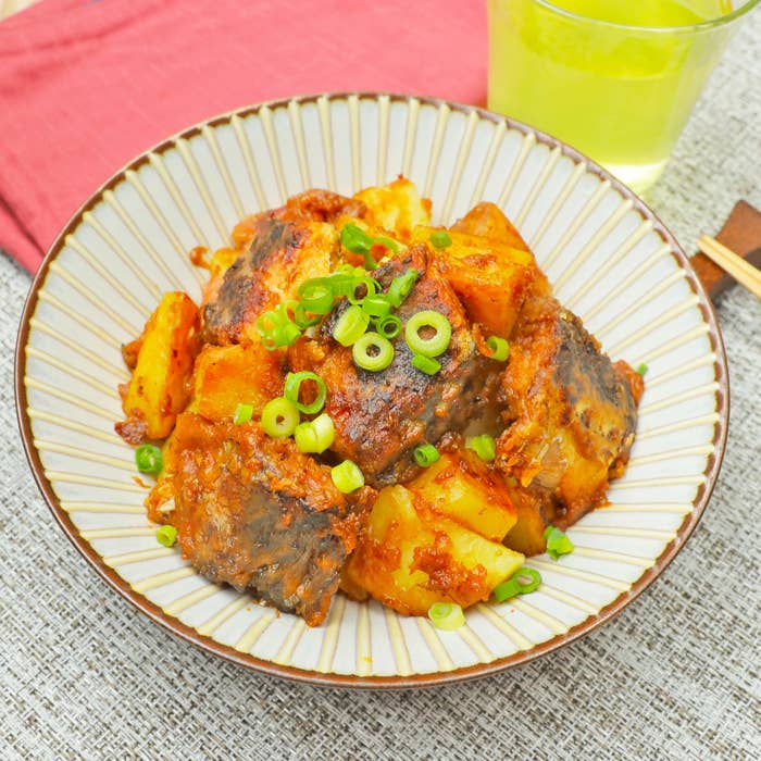 小皿に盛られたピリ辛のじゃがいもの料理。上には刻んだネギがトッピングされている。背景にグラスが見える。