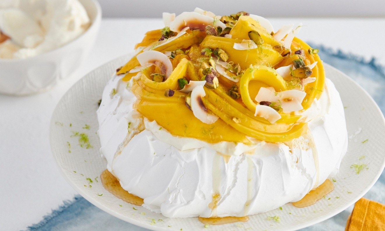 A pavlova dessert topped with whipped cream, mango slices, and crushed pistachios on a white plate