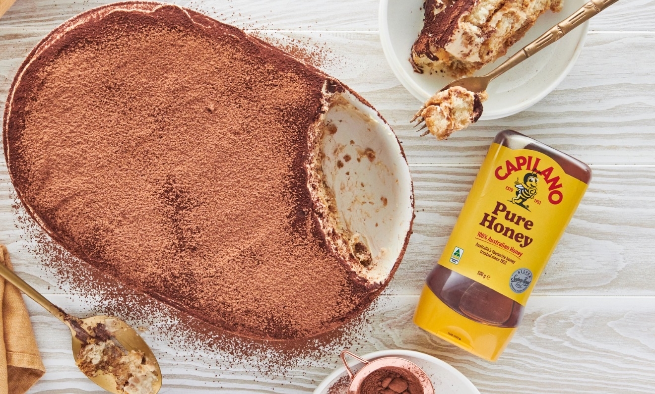 A dish of partially eaten tiramisu, a spoon, a small bowl of tiramisu, a small bowl of cocoa powder, and a bottle of Capilano Pure Honey on a wooden surface