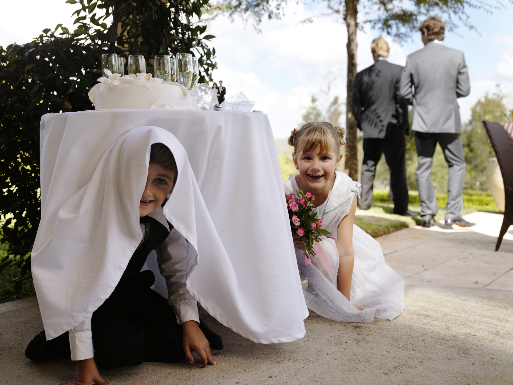 Children play hide-and-seek under a wedding table. Two guests in suits stand in the background