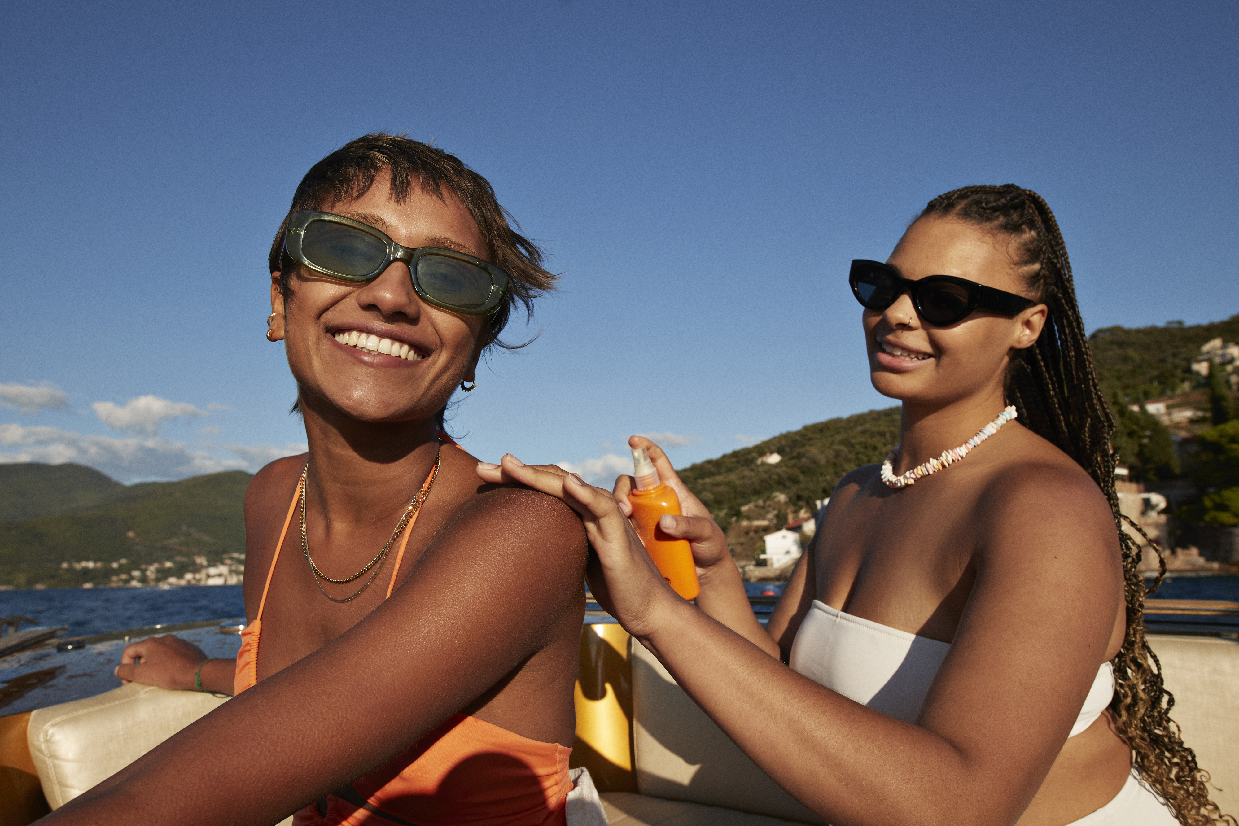 Two women, one with short hair in an orange bikini and sunglasses, the other with braids wearing a white bikini top and sunglasses, applying sunscreen on a boat