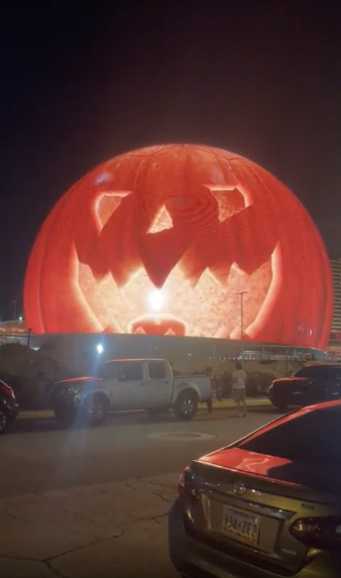 Gigantic pumpkin jack-o'-lantern structure illuminated at night, surrounded by parked cars and a few people