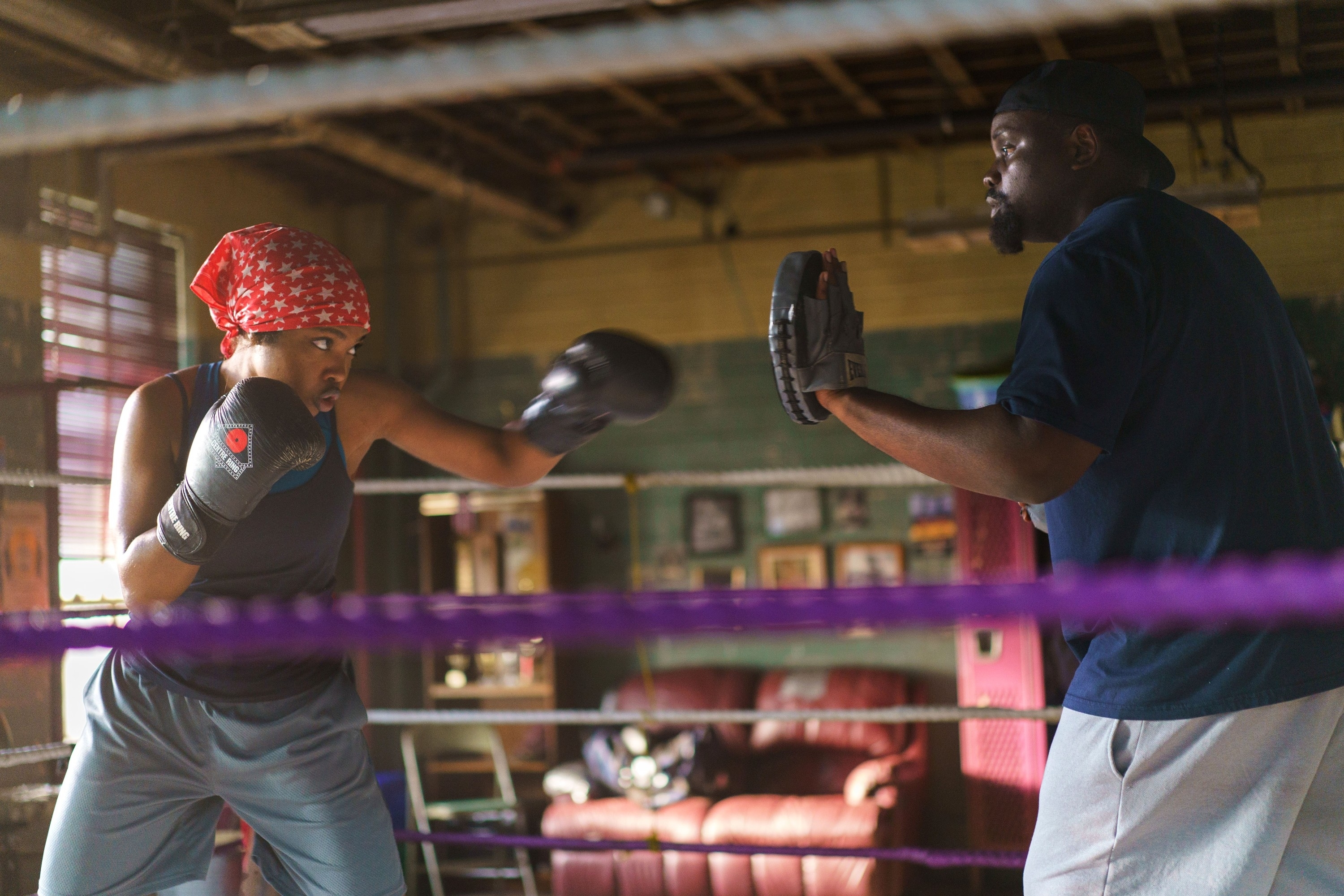 Two people are in a boxing ring. The person on the left, wearing a red bandana, is sparring with the person on the right, holding focus mitts