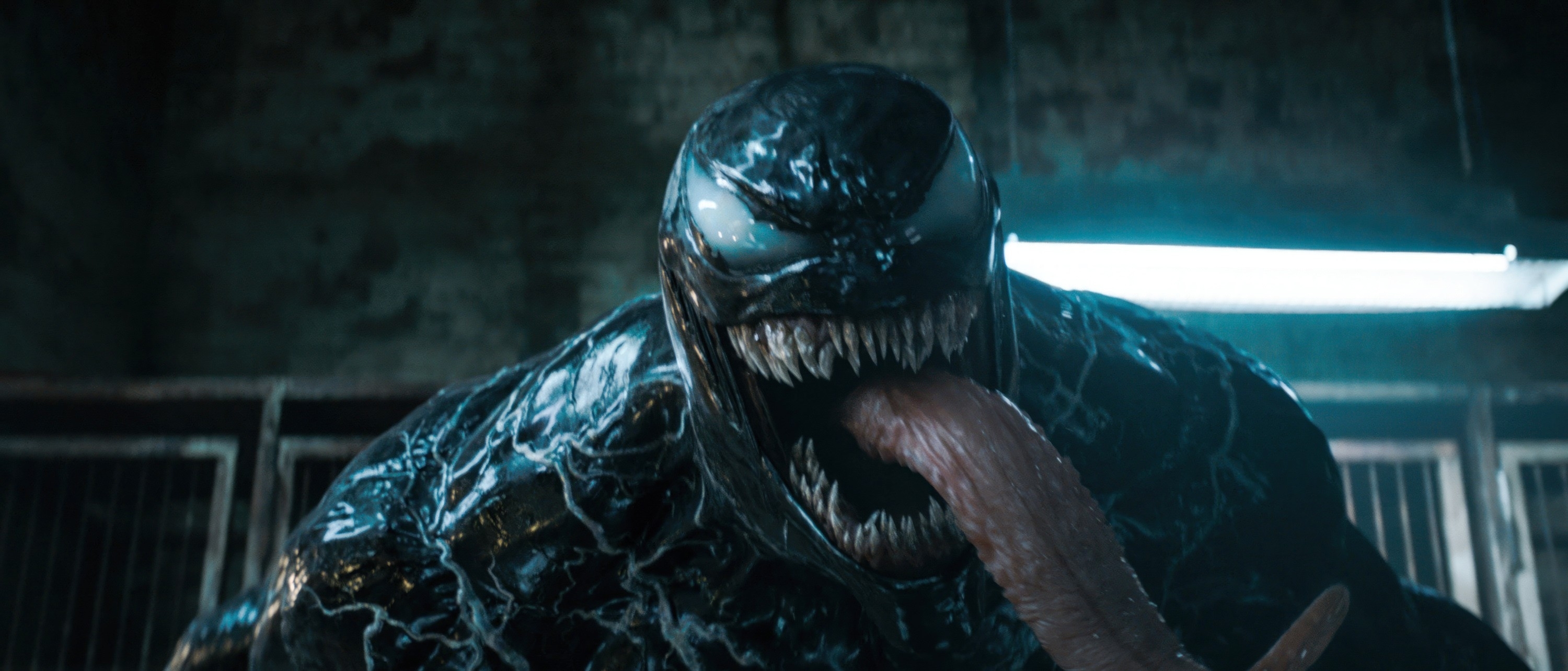 Venom, a character from Marvel Comics, in a dimly lit industrial setting with his mouth open and tongue extended