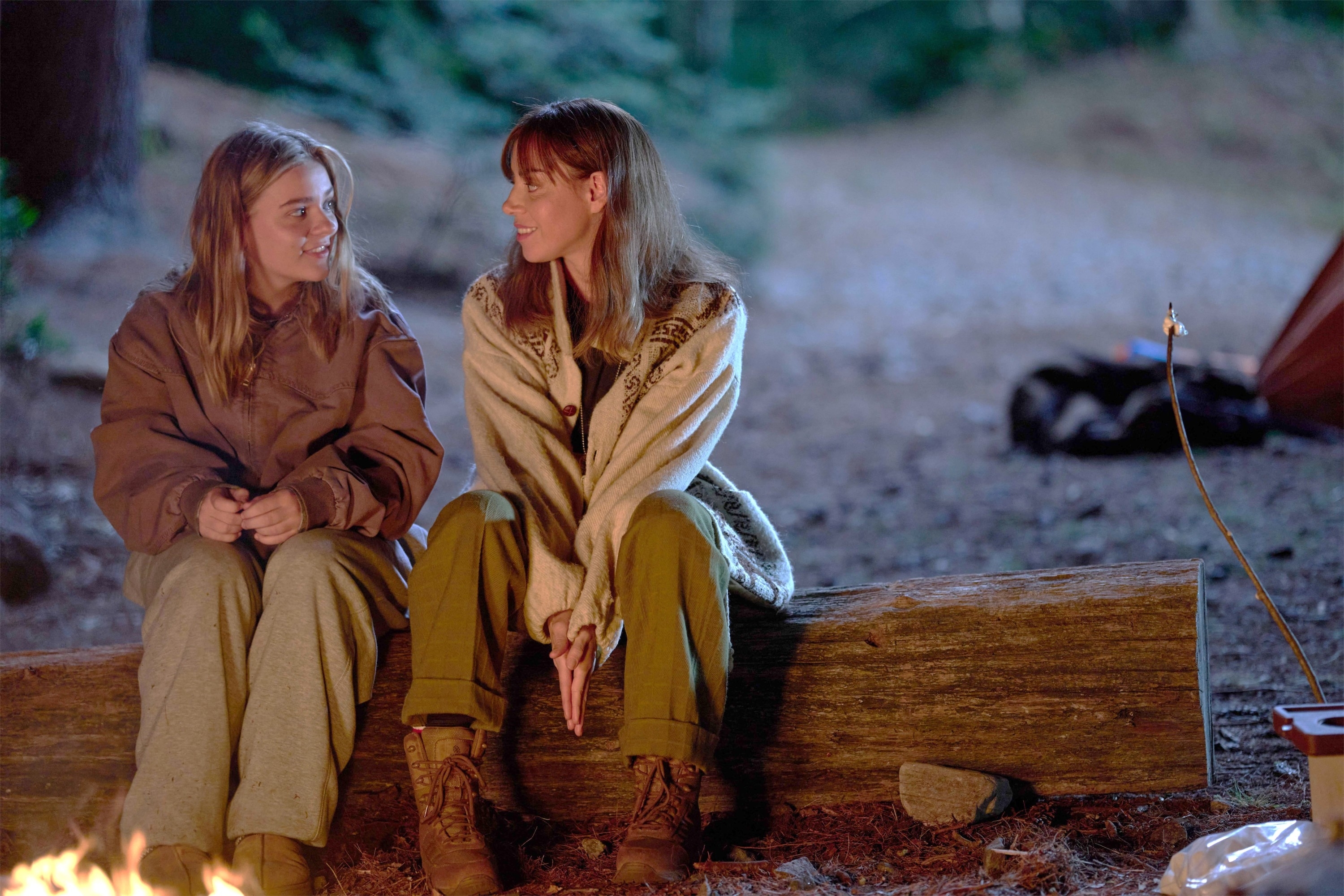 Mary-Kate Olsen and Ashley Olsen sit on a log by a campfire in cozy, casual outdoor clothing, engaging in conversation