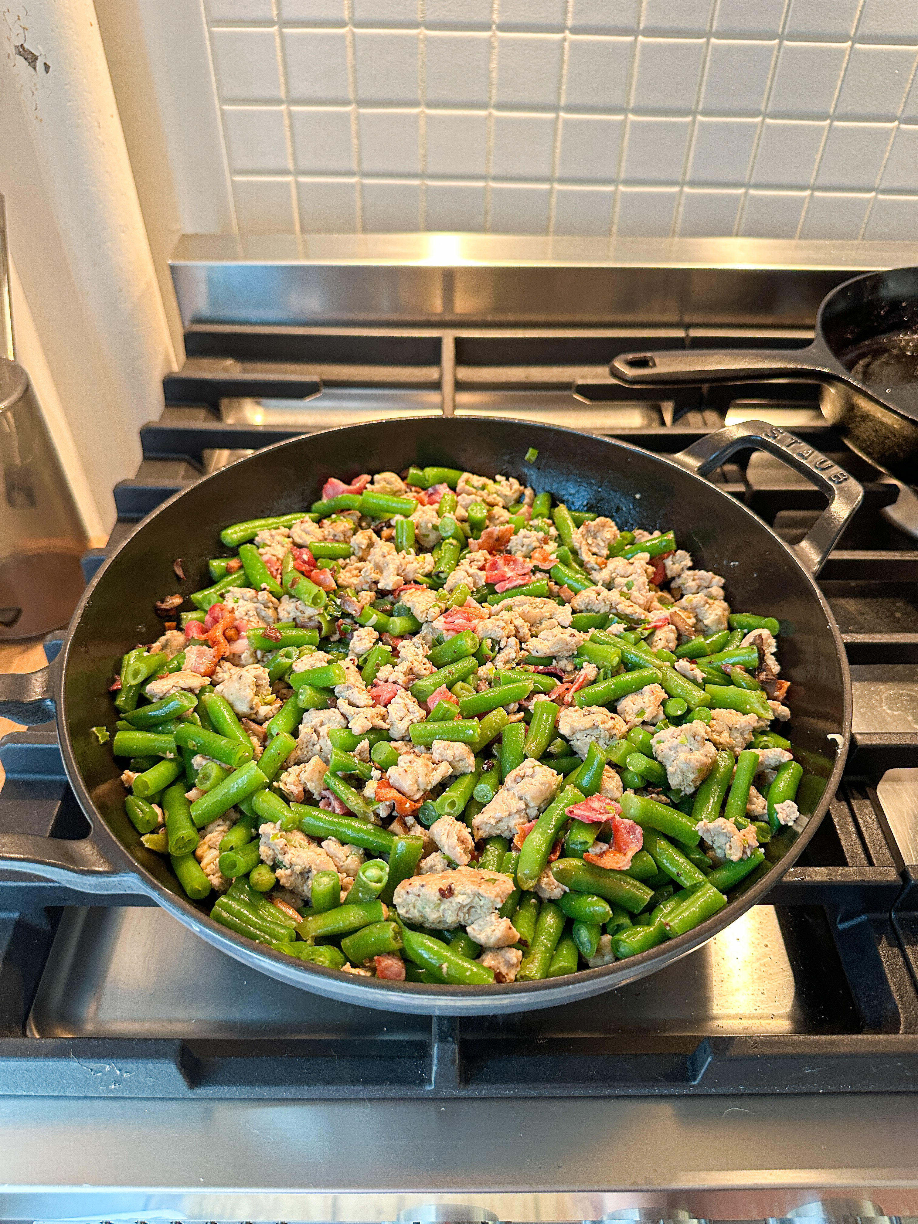 A skillet on a stovetop filled with cooked ground turkey, green beans, and pieces of bacon