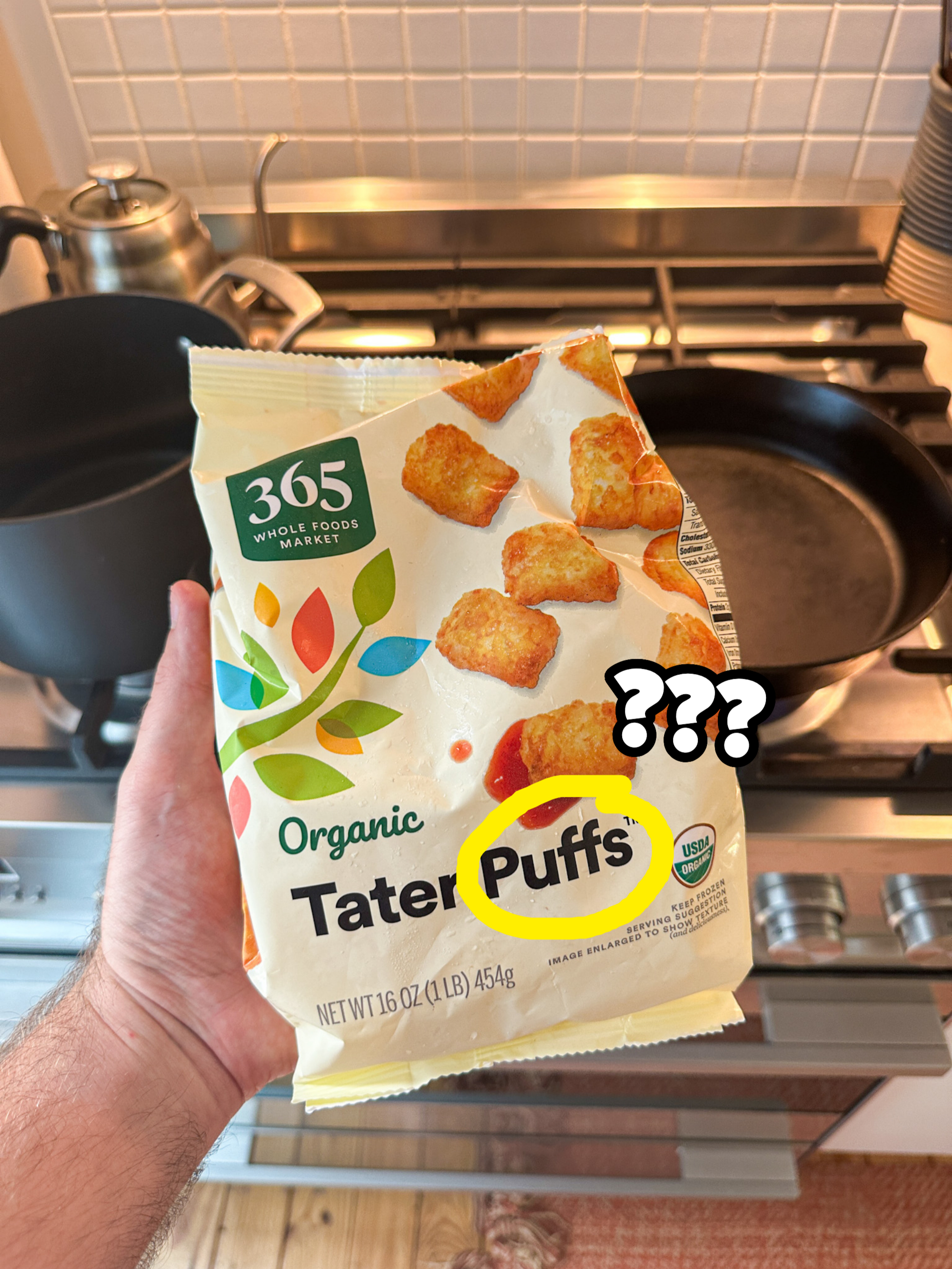Person holding a bag of 365 Whole Foods Market Organic Tater Puffs in a kitchen with a stovetop in the background