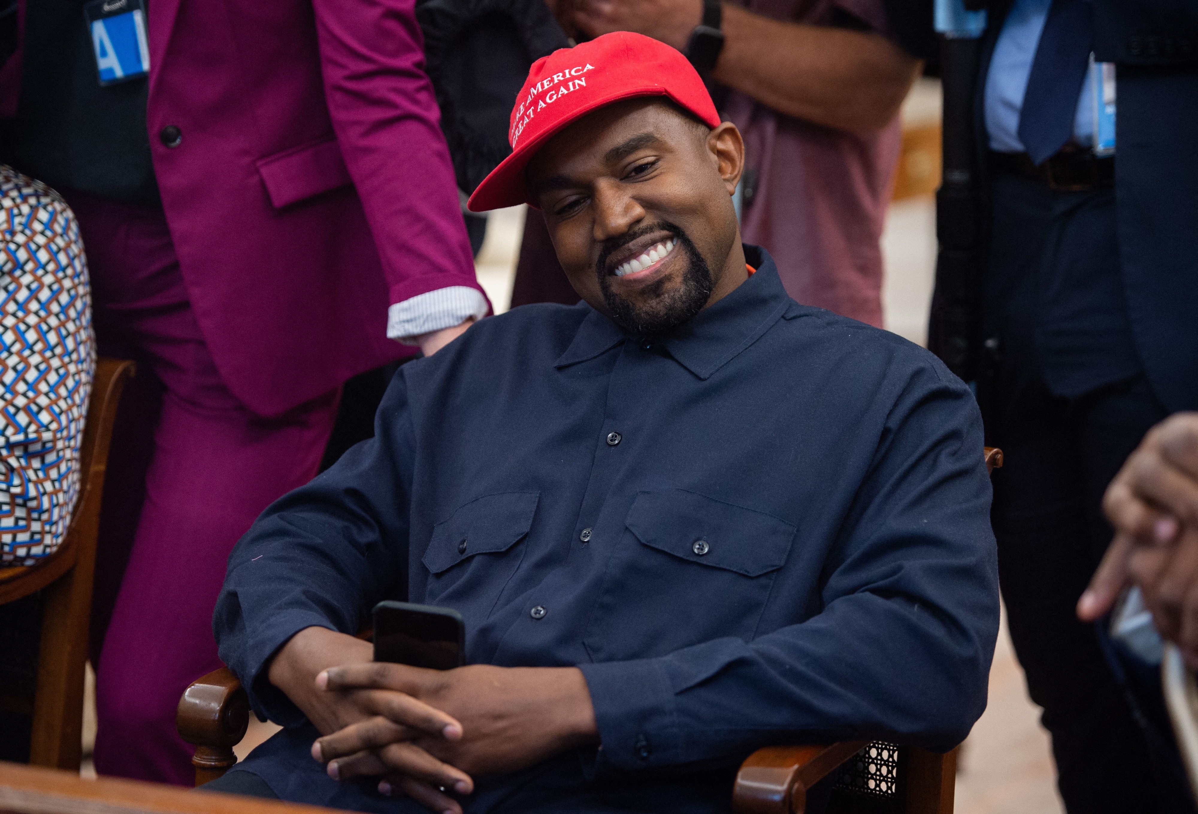 Kanye West, wearing a baseball cap and dark button-down shirt, smiles while looking at his phone. Several people, including one in a pink suit, are around him