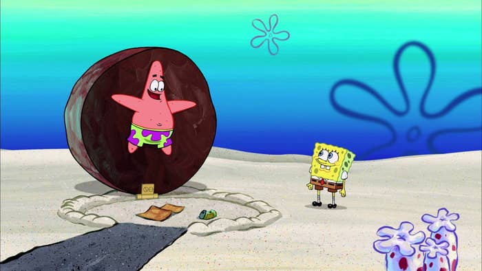 Patrick Star stands in front of a large chocolate cookie, smiling. SpongeBob SquarePants watches nearby on a sandy beach