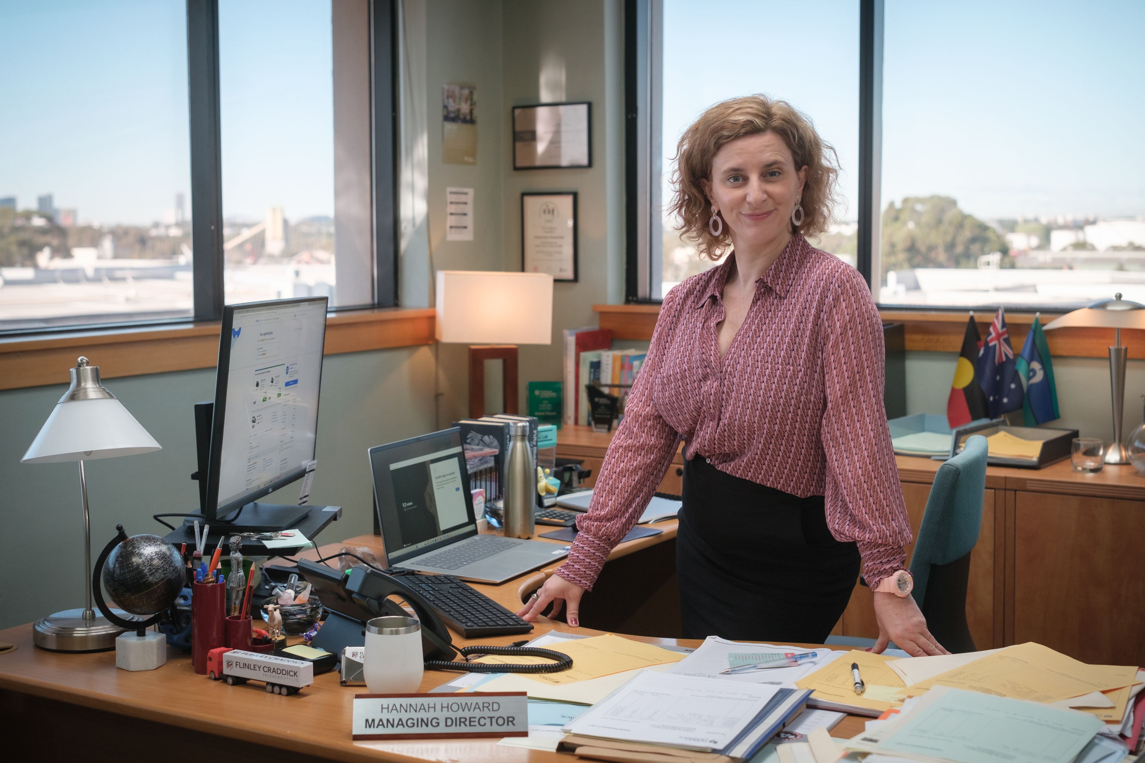 Hannah Howard stands confidently in her office, which has a desk cluttered with papers, a computer, and a globe. A nameplate reads "Hannah Howard, Managing Director."