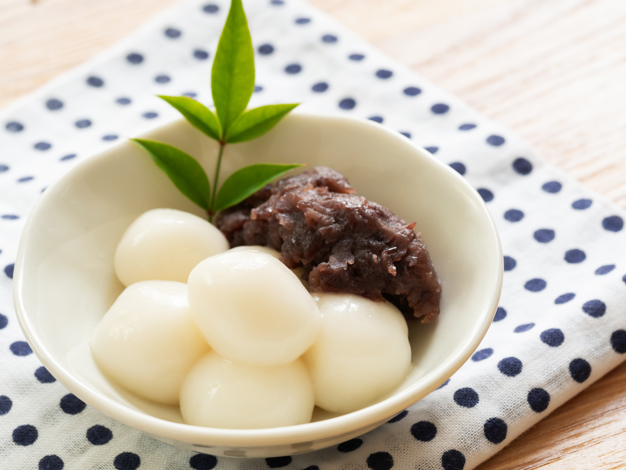 白いお団子が数個に団子の上にのっている小皿に、緑色の葉とあんこが添えられている。背景には水玉模様の布。
