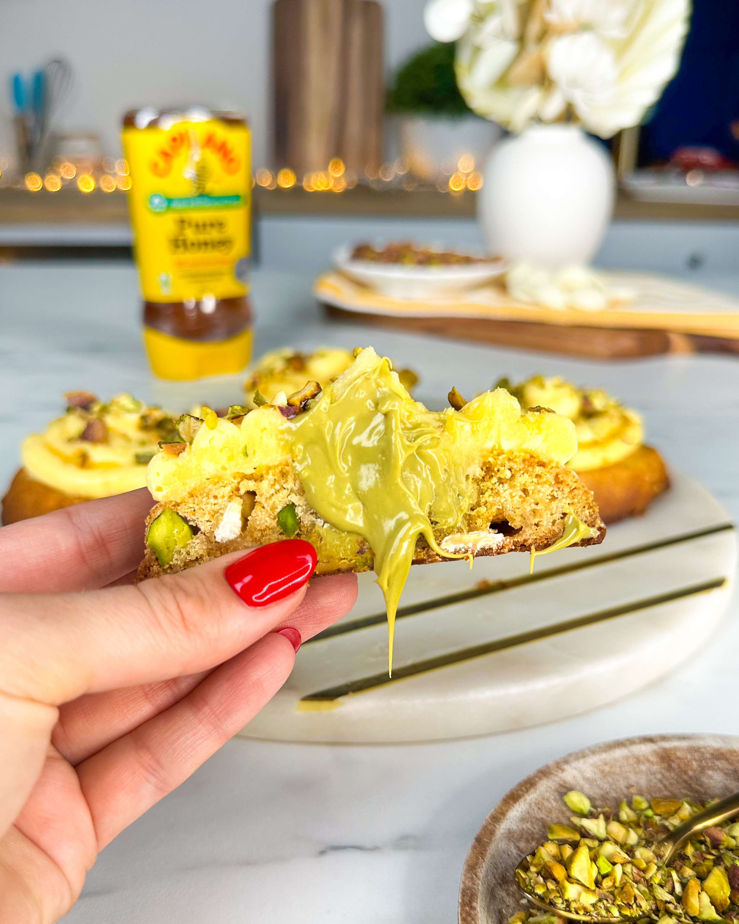 Close-up of hand holding a slice of bread topped with pistachios and yellow spread, with a bottle of honey and other food items in the background on a table