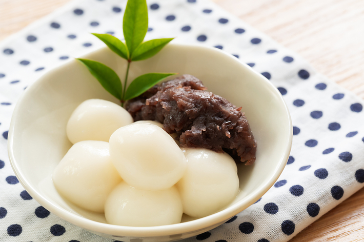 白いお団子が数個に団子の上にのっている小皿に、緑色の葉とあんこが添えられている。背景には水玉模様の布。