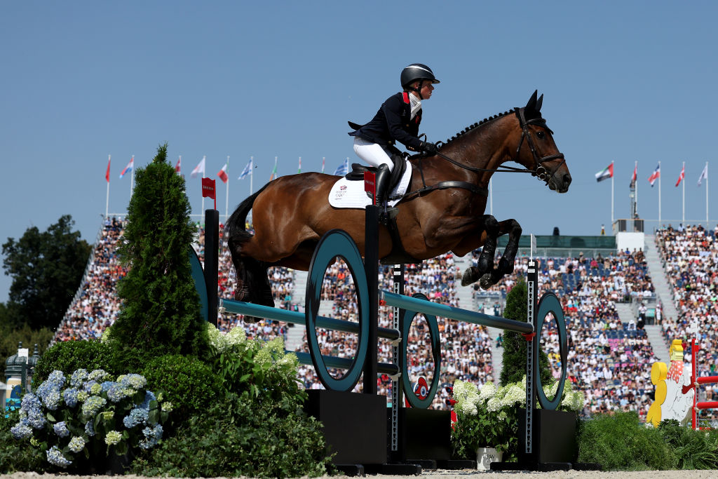 Equestrian Charlotte Dujardin jumps a hurdle on horseback during a competition, with a large audience in the stadium background