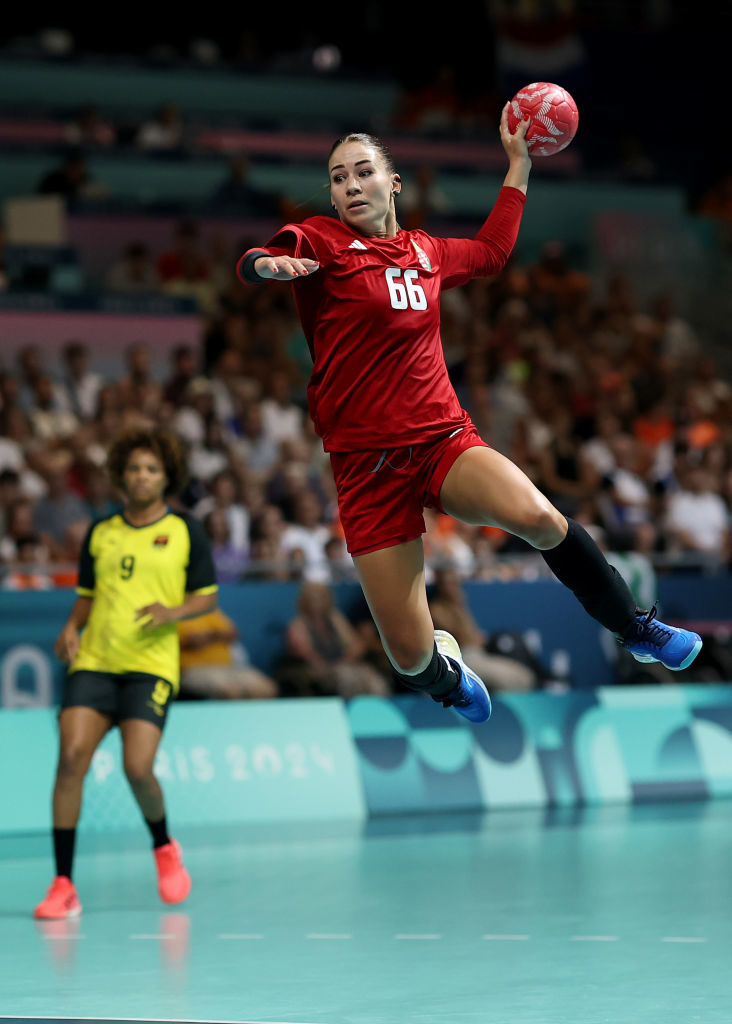 Two female handball players in action during a game. Player in red jersey number 66 is jumping to throw the ball, while player in yellow jersey number 9 looks on