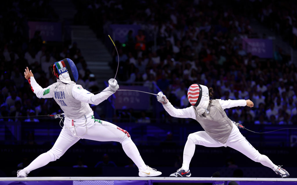 Two fencers in action during a match, identified as Pozdziulkina and Prescod, wear protective gear and face off with foil swords. The background is a packed arena
