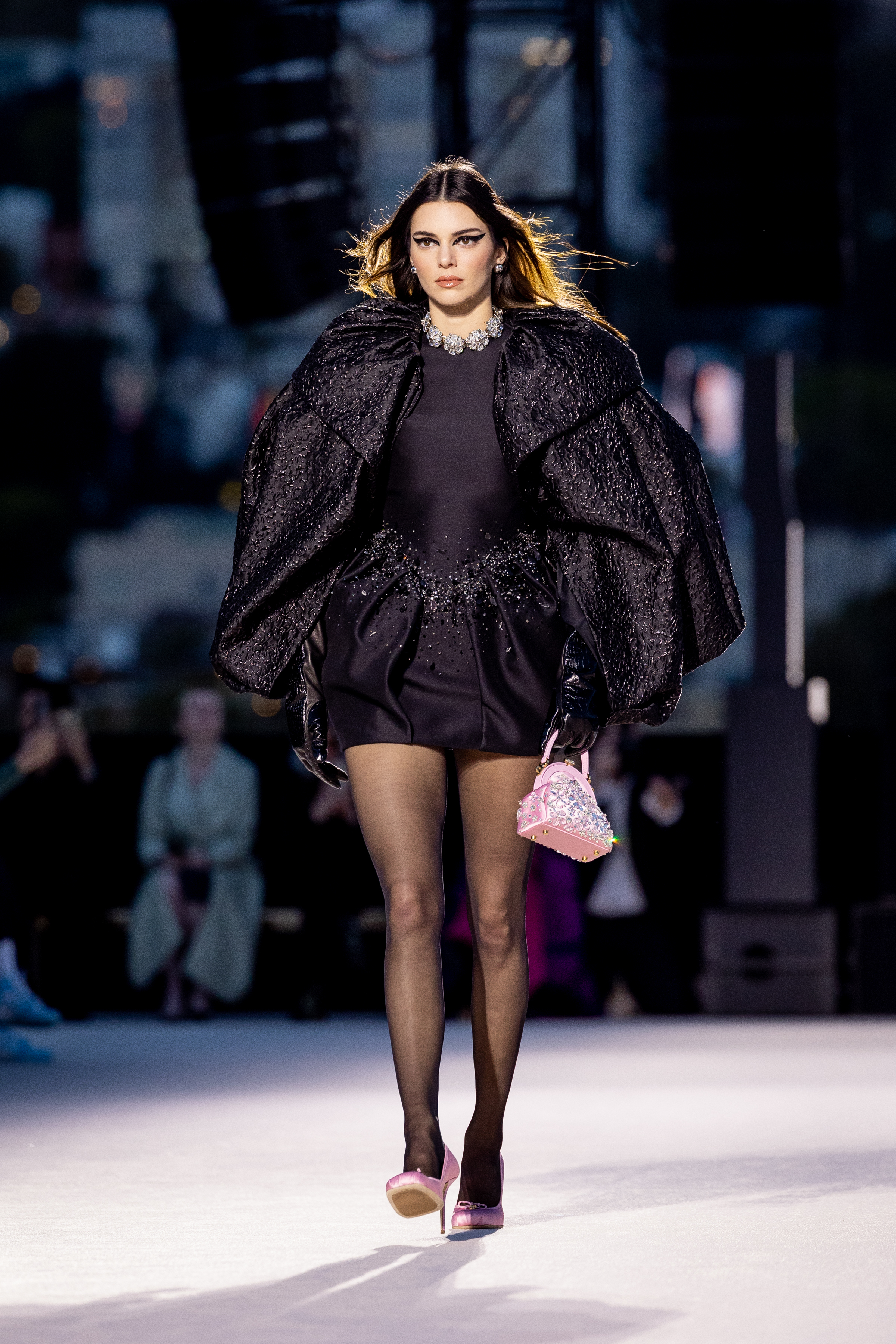 Kendall Jenner walking on a runway wearing a short dark dress with a textured cape, dark gloves, sheer dark tights, pink shoes, and carrying a pink handbag