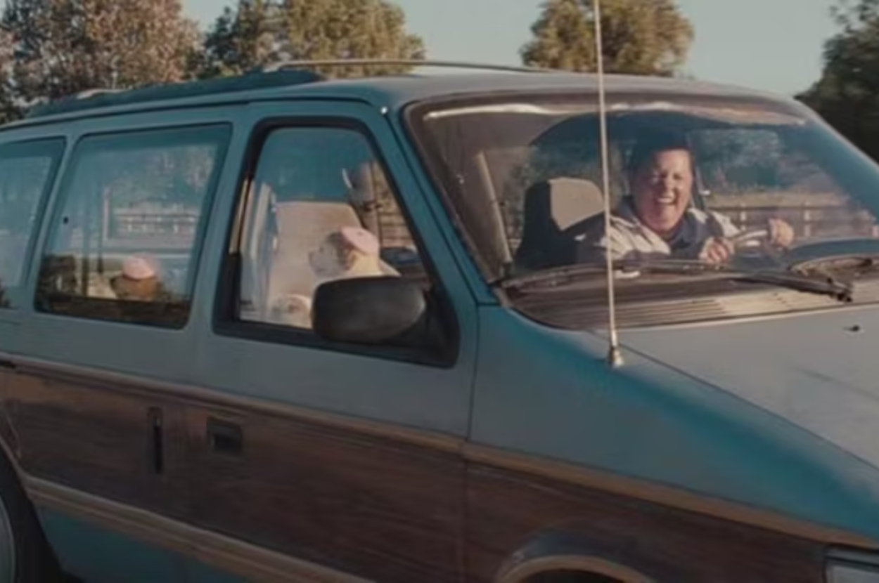 A woman dressed casually is joyfully driving a minivan with a dog in the passenger seat. Trees are seen in the background