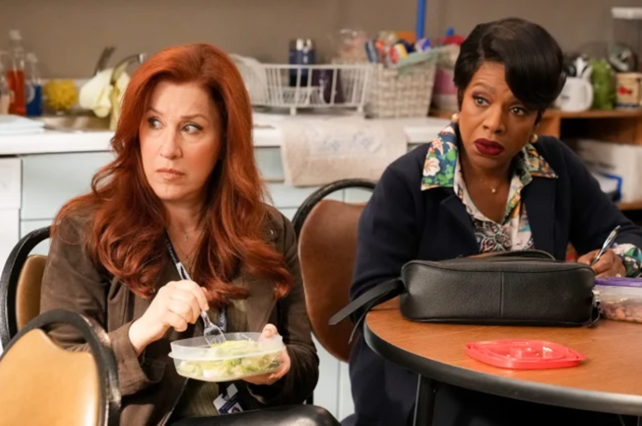 Annie Potts and Sheryl Lee Ralph sit in a break room. Annie is holding a plastic container and fork, while Sheryl looks on