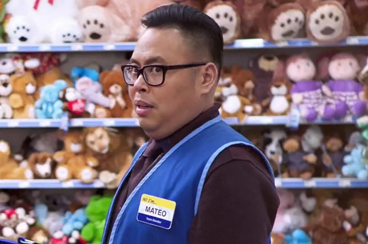 Nico Santos as Mateo wearing a store employee vest with a nametag in front of a shelf filled with plush toys