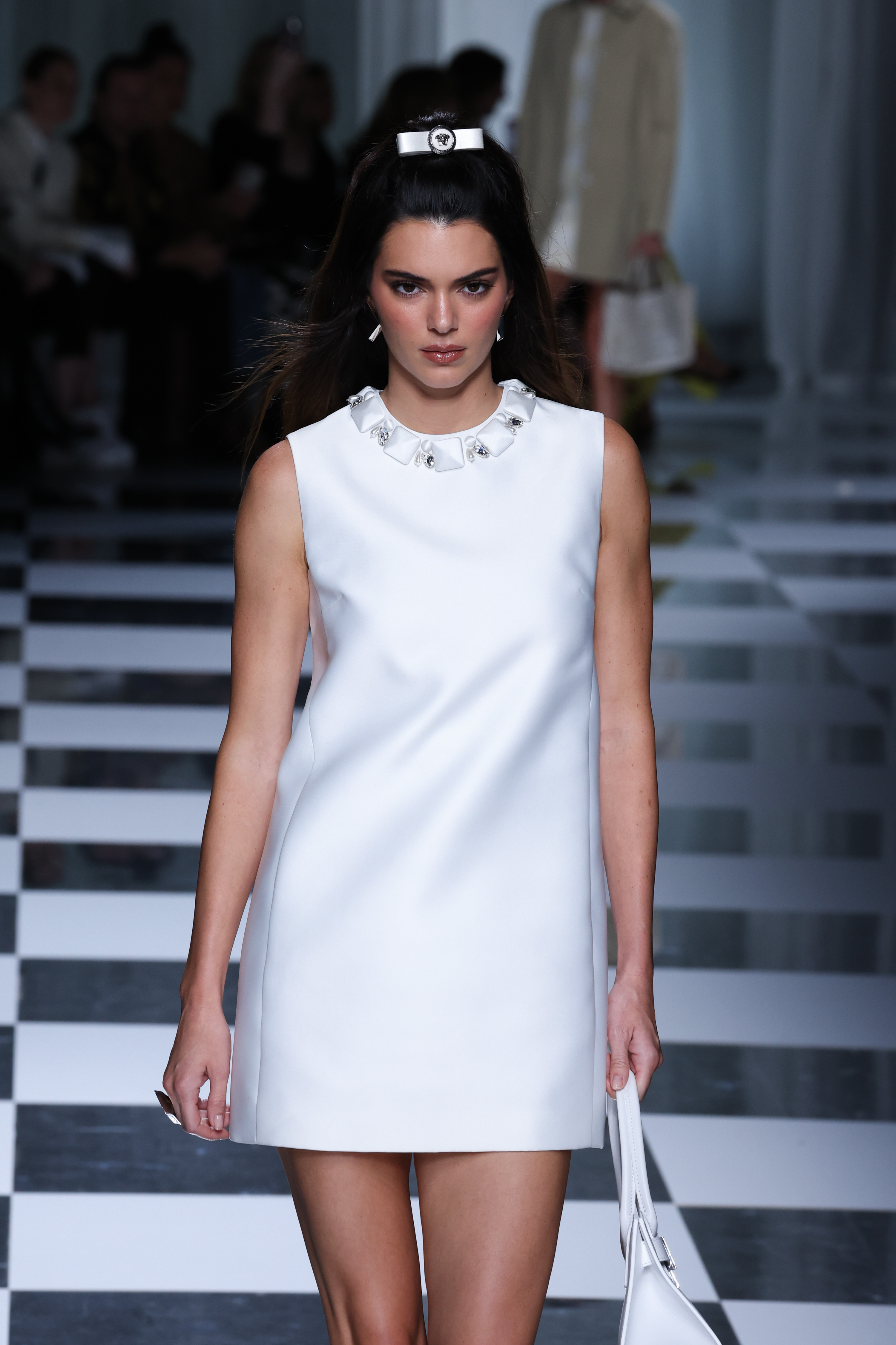 Kendall Jenner walks down the runway in a sleeveless, above-the-knee dress with a high neckline, and an embellishment at the collar, carrying a handbag