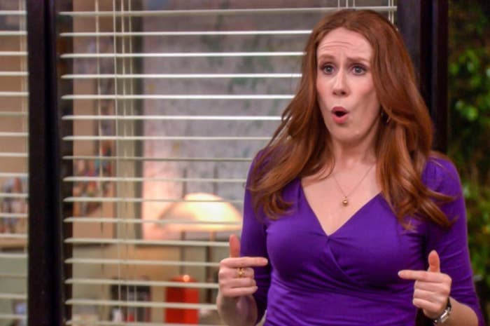 A woman with long hair stands indoors wearing a wrap-style top and appears to be speaking or reacting emotionally