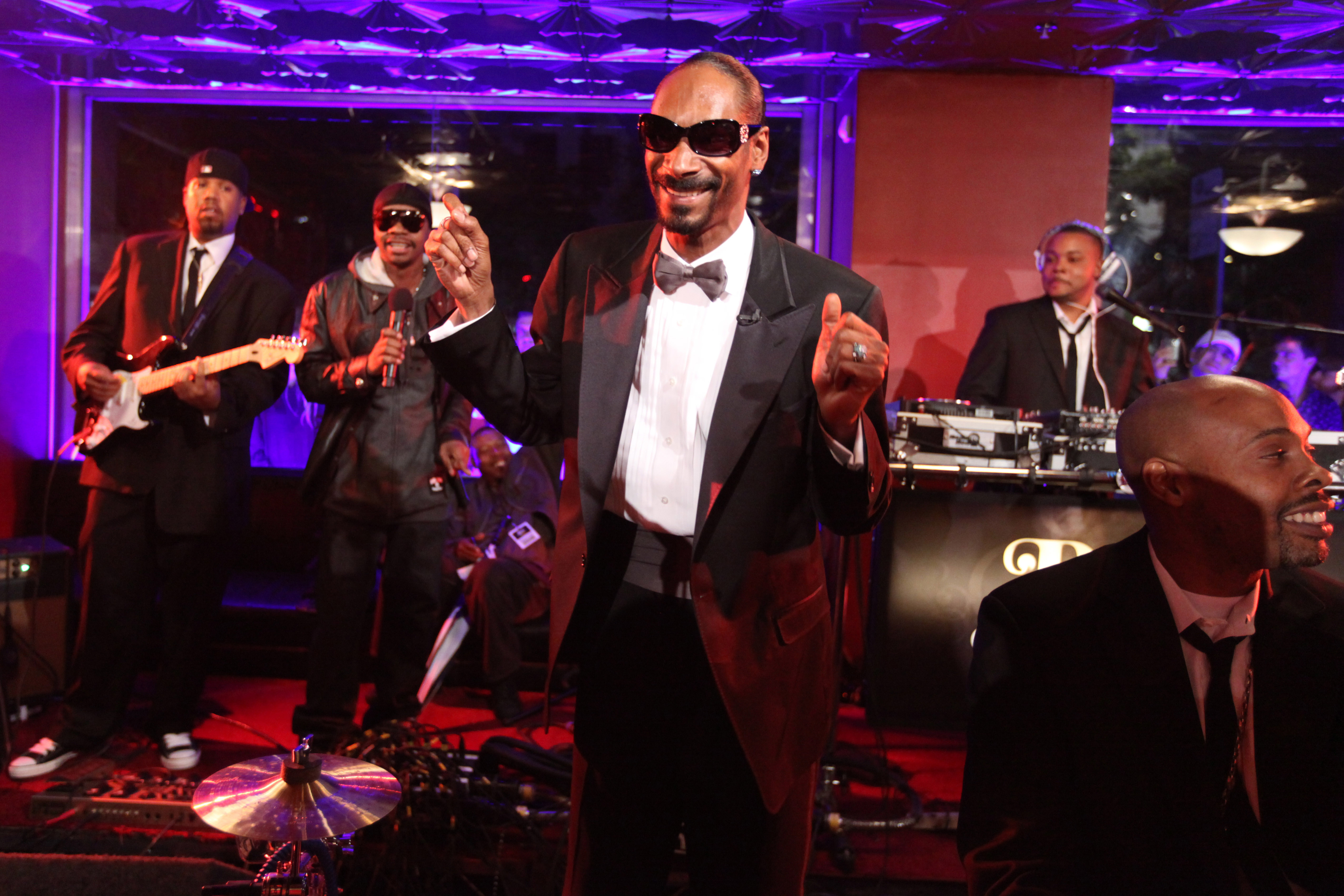 Snoop Dogg performs on stage wearing a tuxedo and sunglasses, accompanied by his band members playing instruments