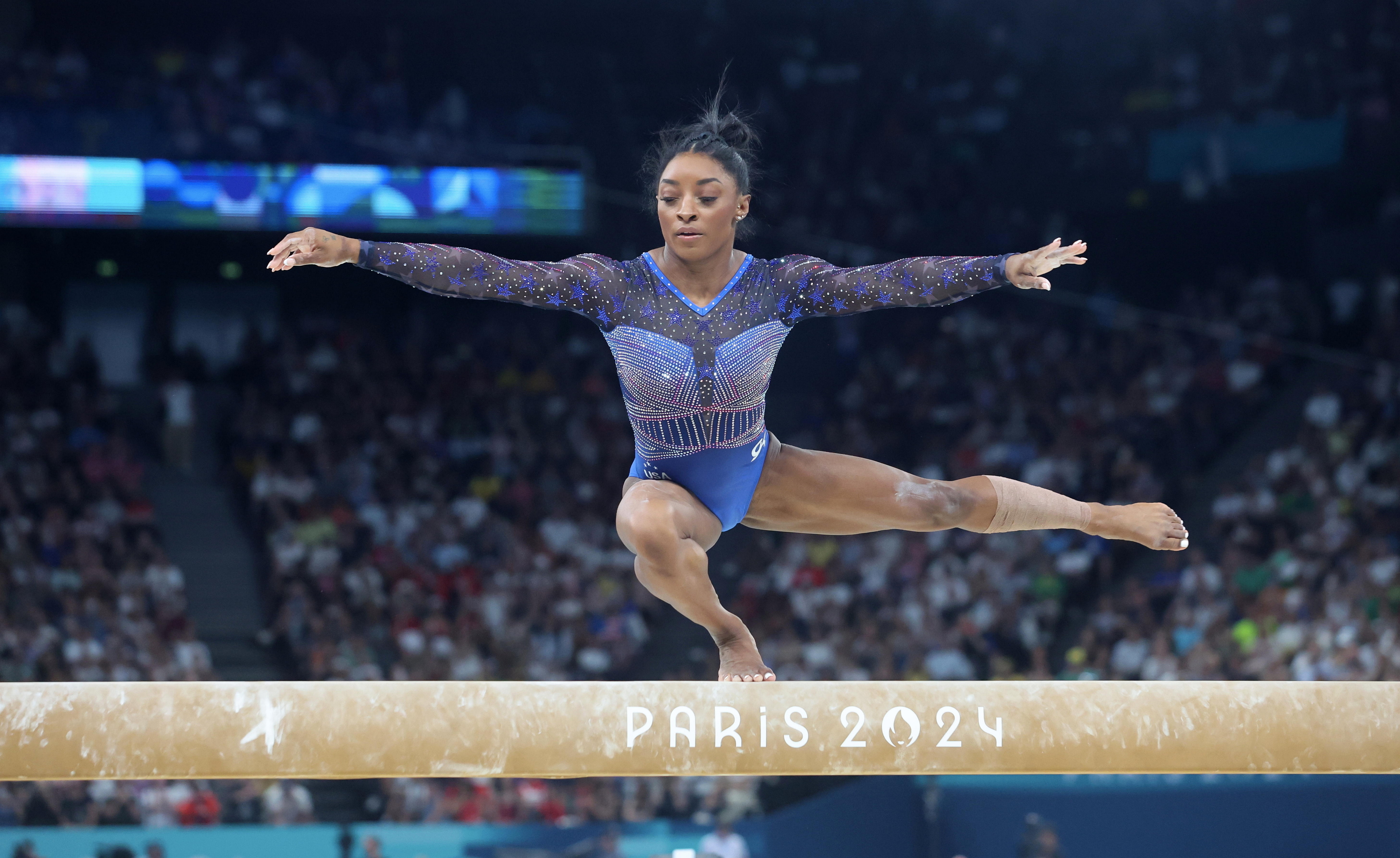 Simone Biles performing a one-legged spin, known as a full turn, on the balance beam at the Paris 2024 gymnastics event, with a focused crowd in the background