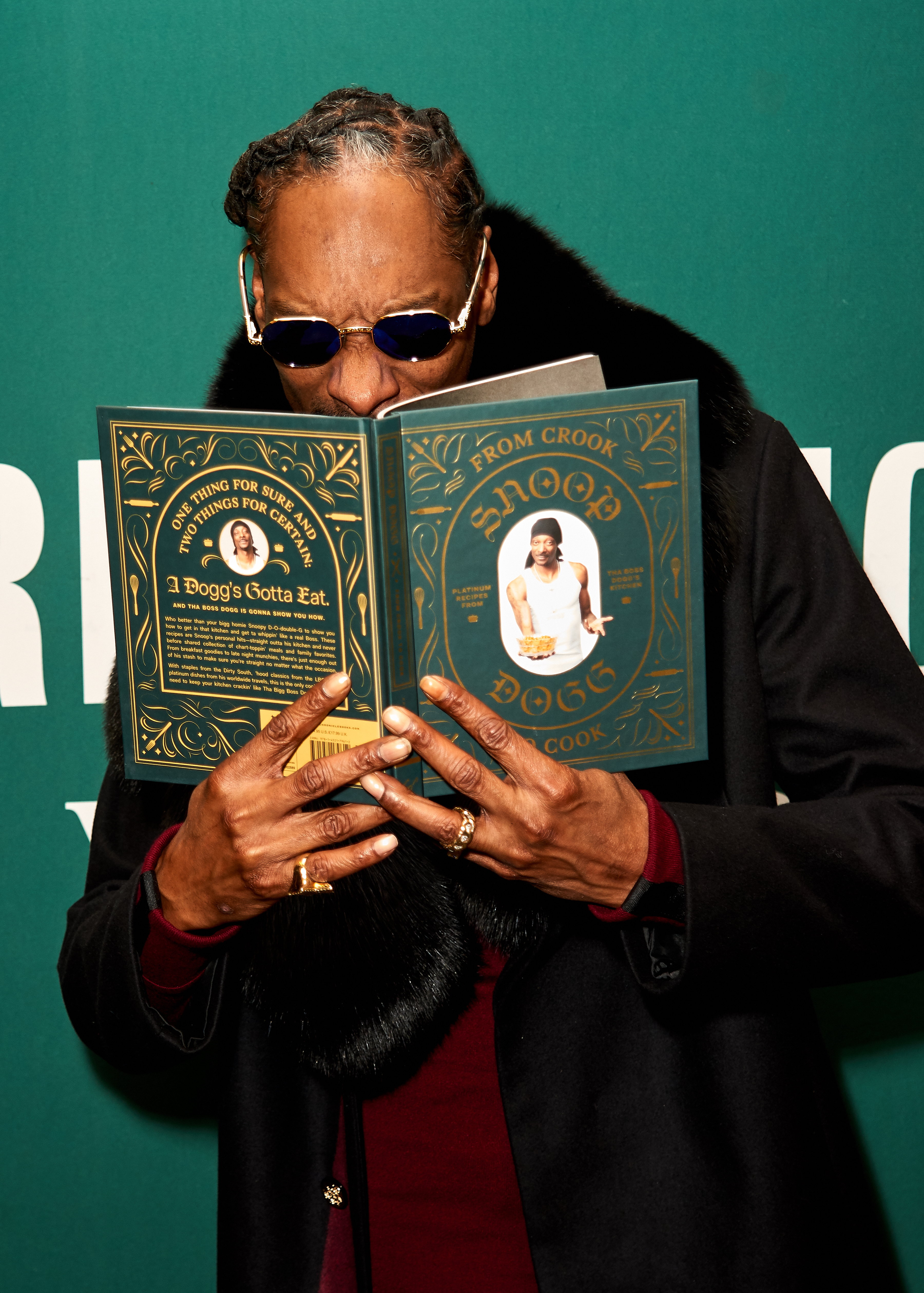Snoop Dogg stands reading his book &quot;From Crook to Cook,&quot; wearing dark sunglasses and a black fur-trimmed jacket