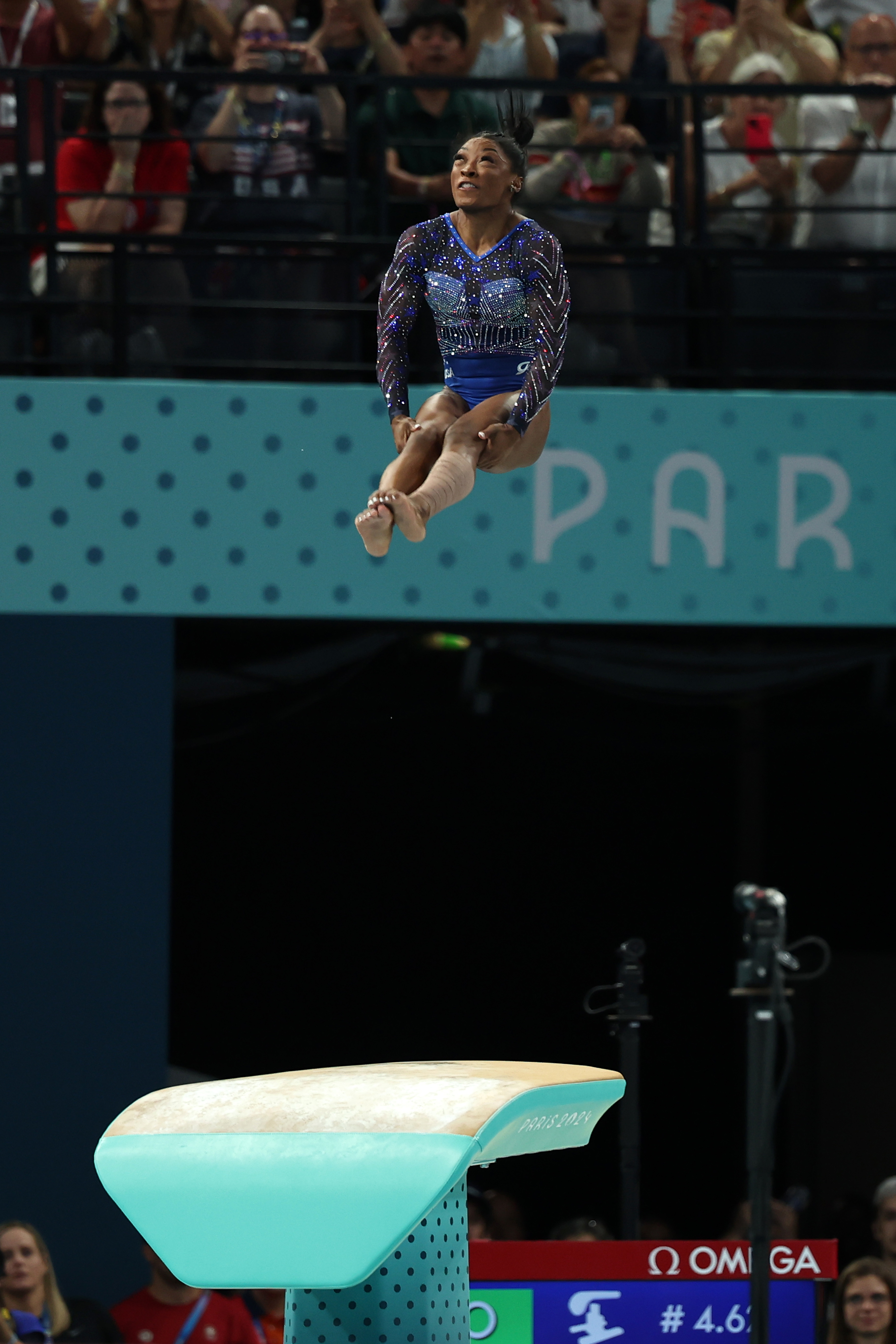 Simone Biles performs a high jump off the vault during a gymnastics competition, surrounded by a cheering audience
