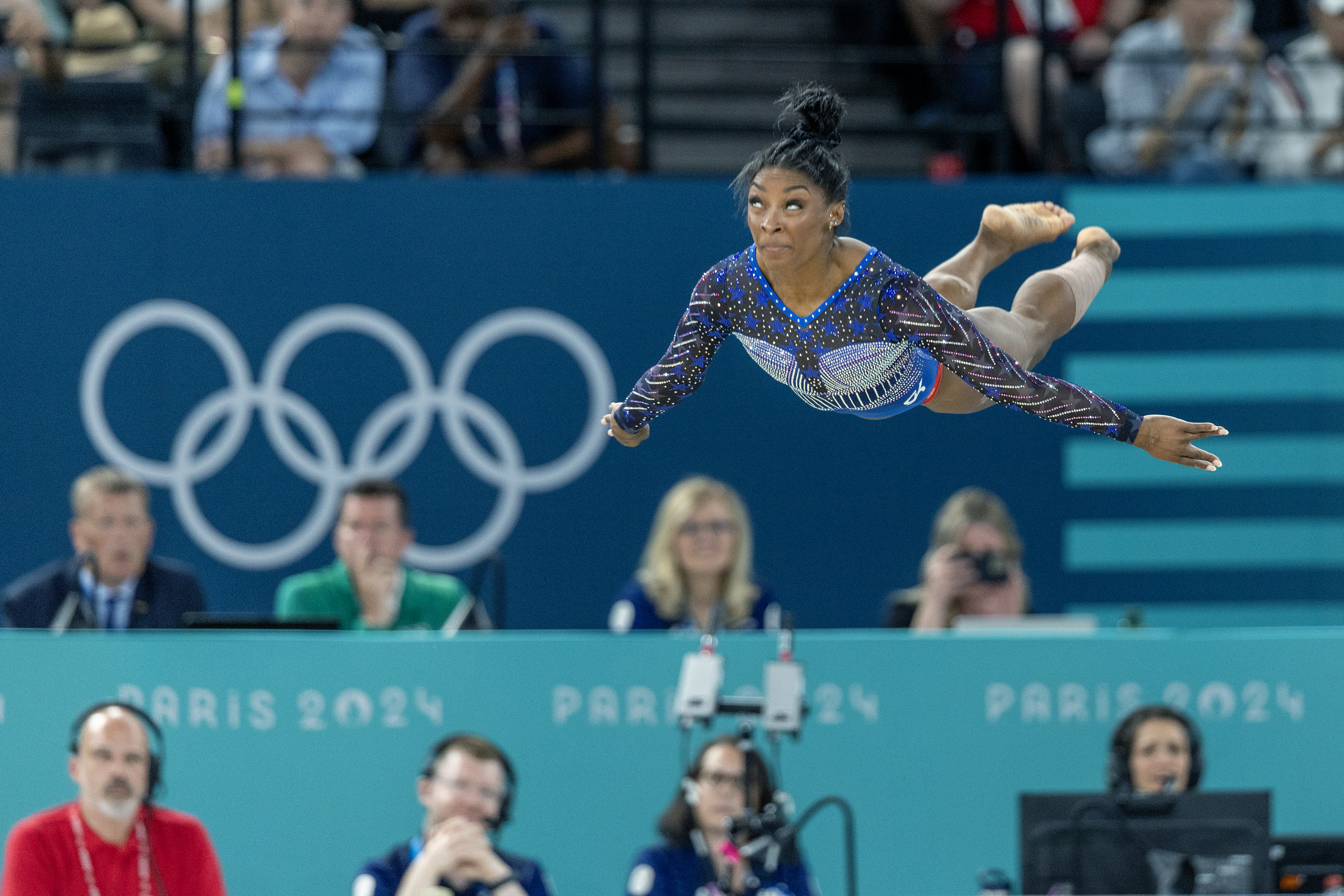 Simone Biles performing a gymnastics routine at the Paris 2024 Olympics, mid-air in an arena filled with spectators