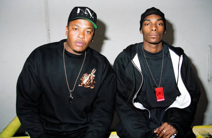 Dr. Dre and Snoop Dogg pose together wearing black sweatshirts and baseball caps, with casual accessories, against a plain background