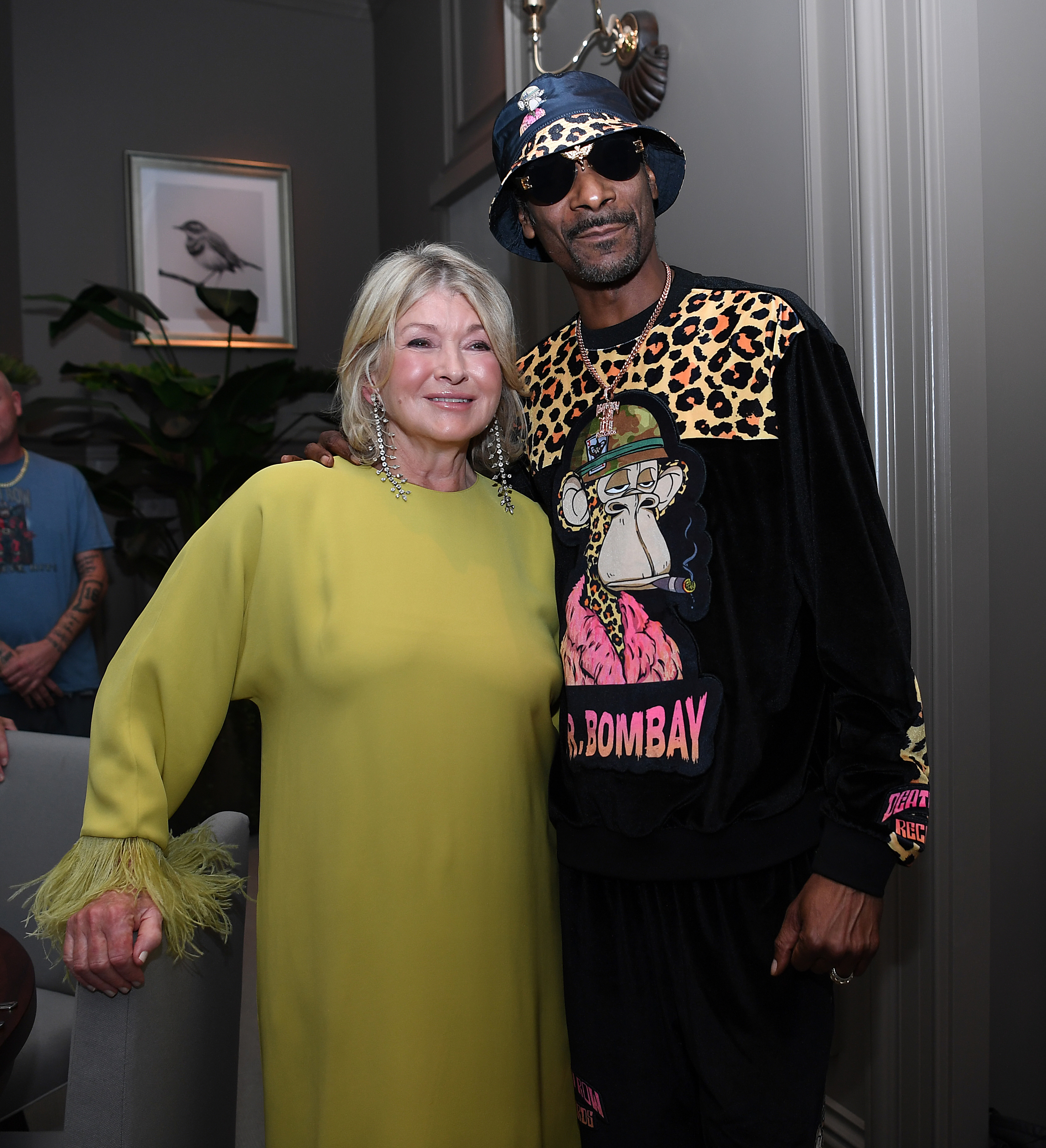 Martha Stewart and Snoop Dogg pose together indoors. Martha wears a flowing dress with feathered sleeves, and Snoop wears a bucket hat and outfit with animal print accents