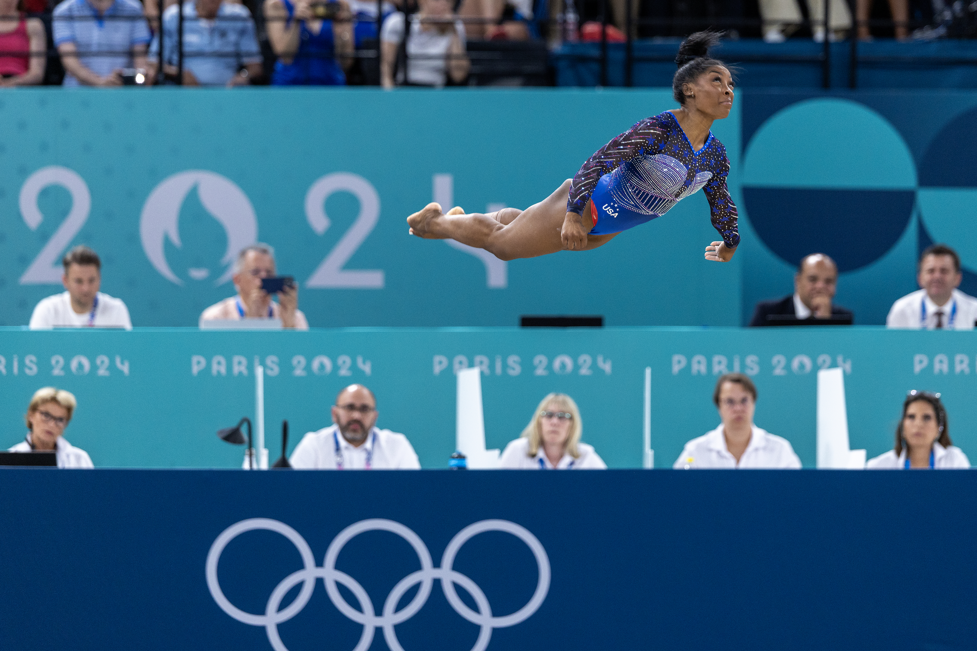 Simone Biles performs a mid-air flip during a gymnastics event at the Paris 2024 Olympics, with judges watching in the background