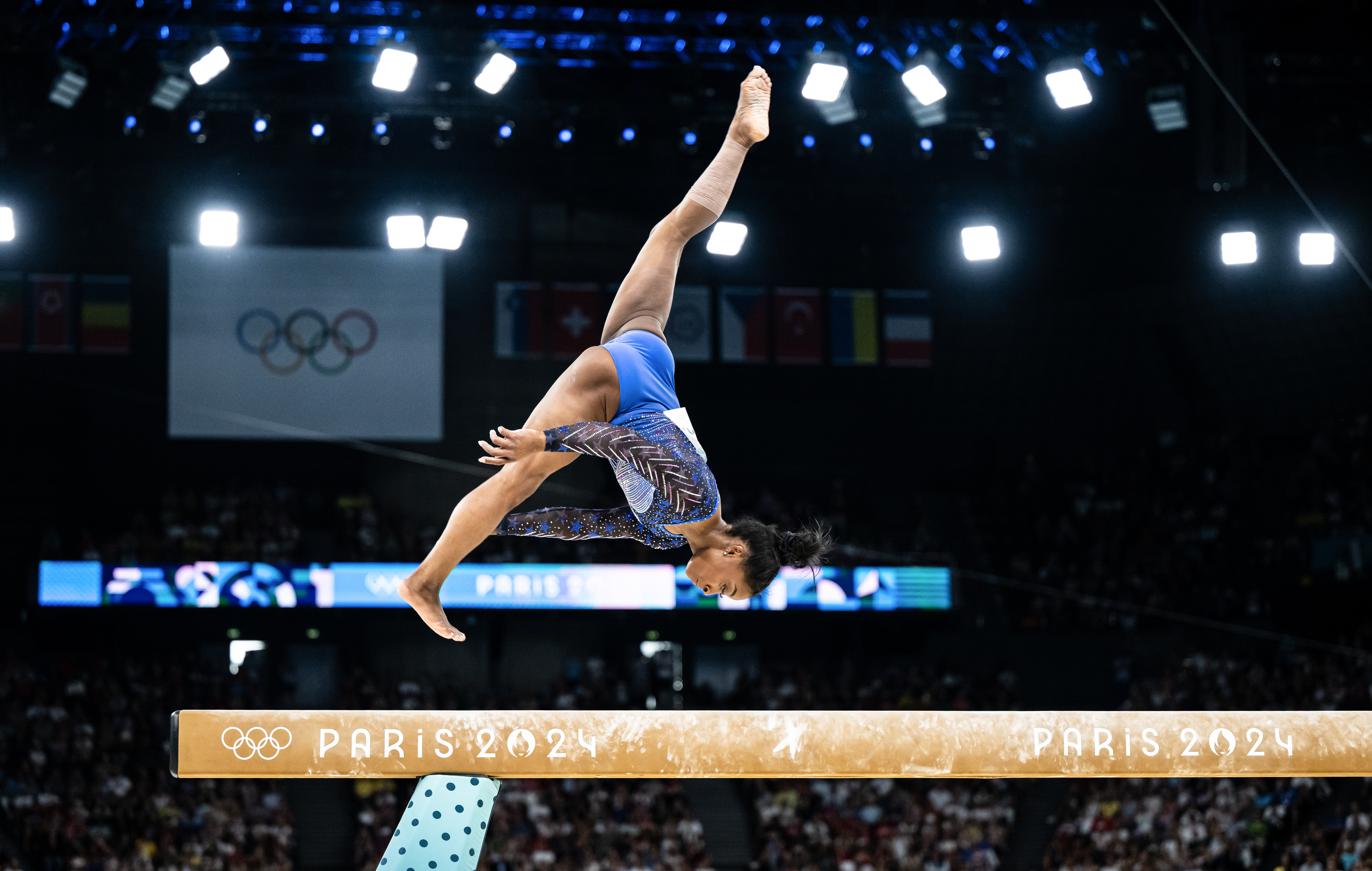 Simone Biles performs an acrobatic routine on the balance beam at the Paris 2024 Olympics, with an arena full of spectators in the background