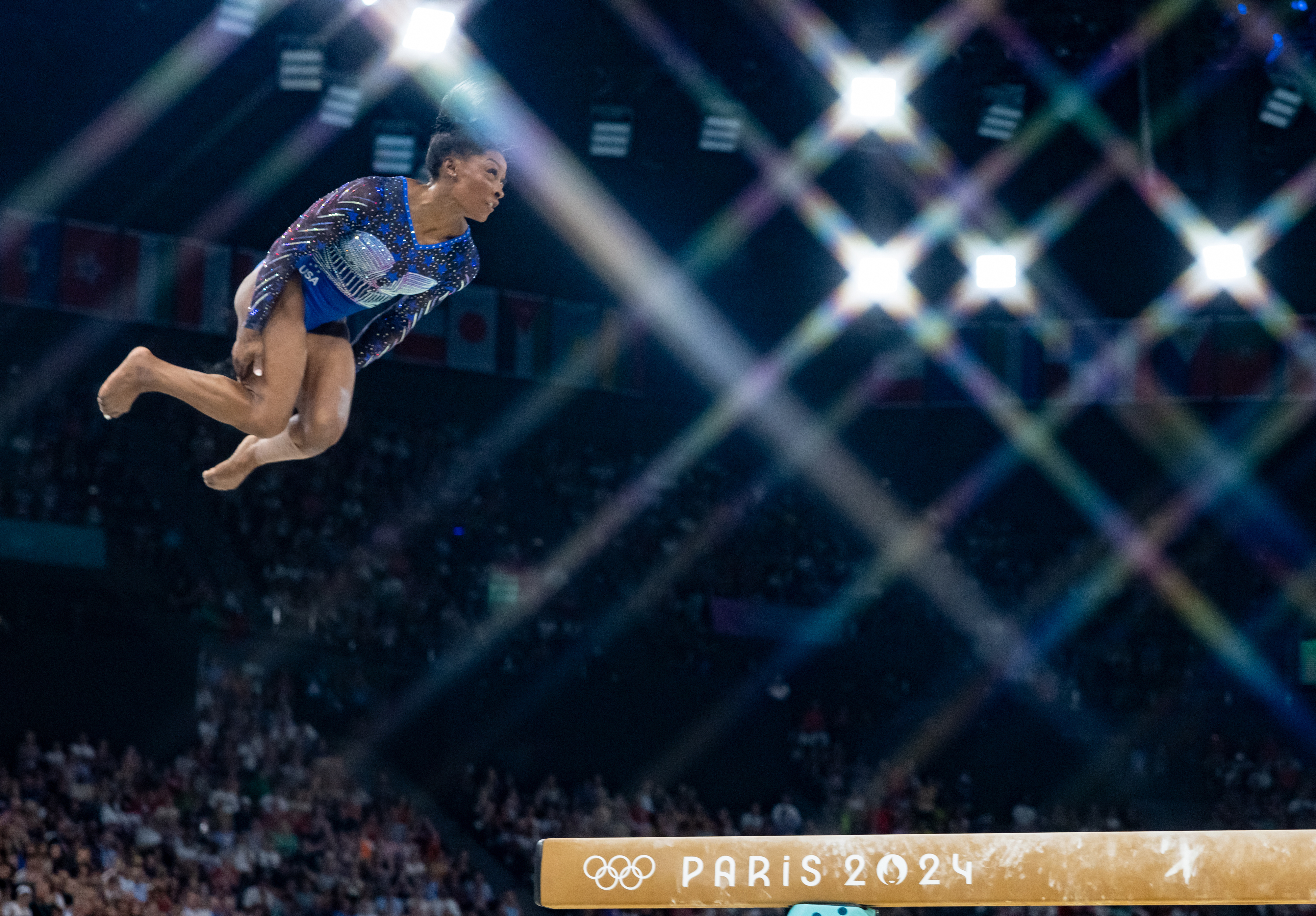 Simone Biles performs a high-flying gymnastics routine during the Paris 2024 Olympics, with the audience and event signage visible in the background