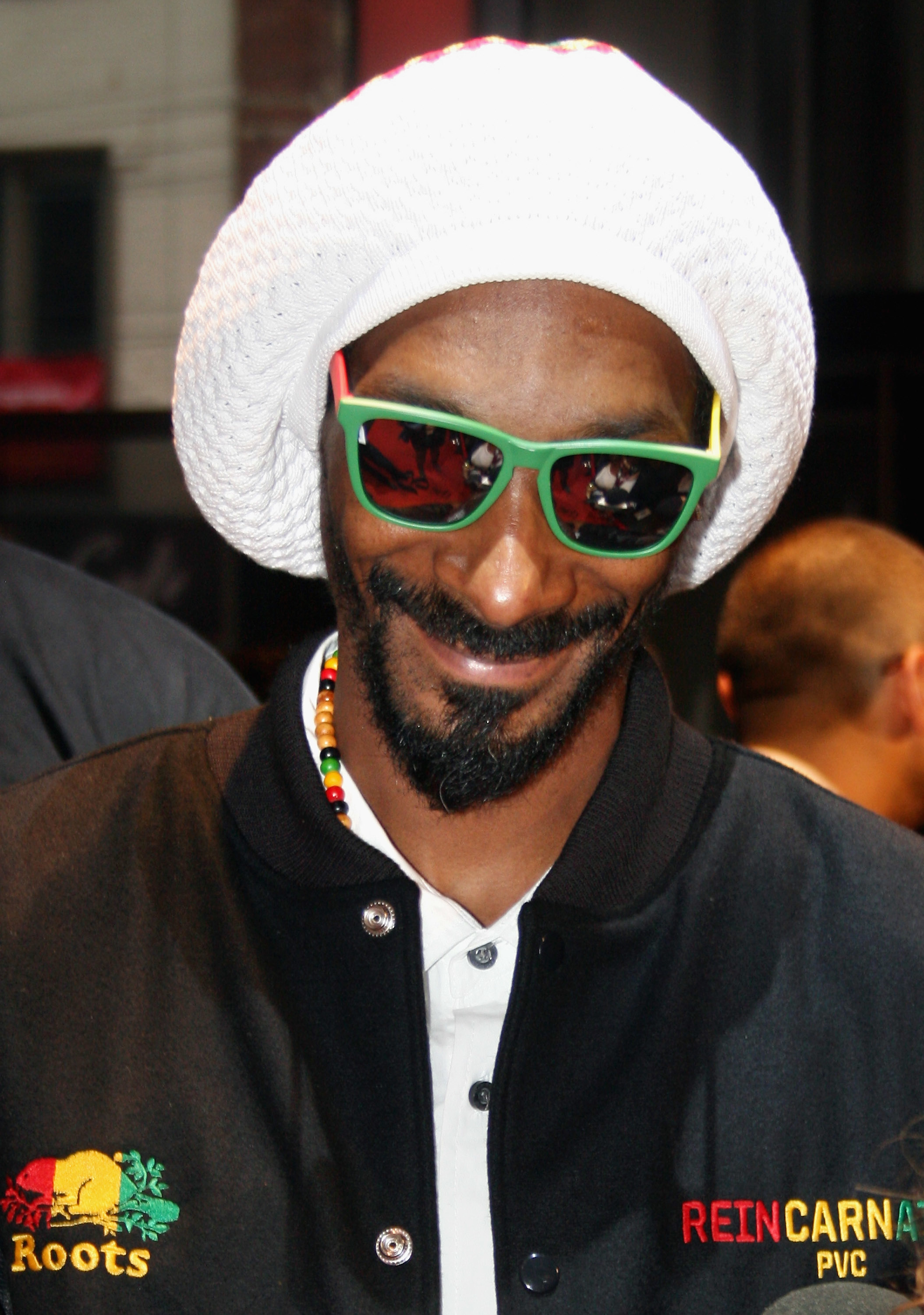 Snoop Dogg wears a white knitted hat, colorful beaded necklace, and green sunglasses, smiling at an event. He has on a black jacket with embroidered patches