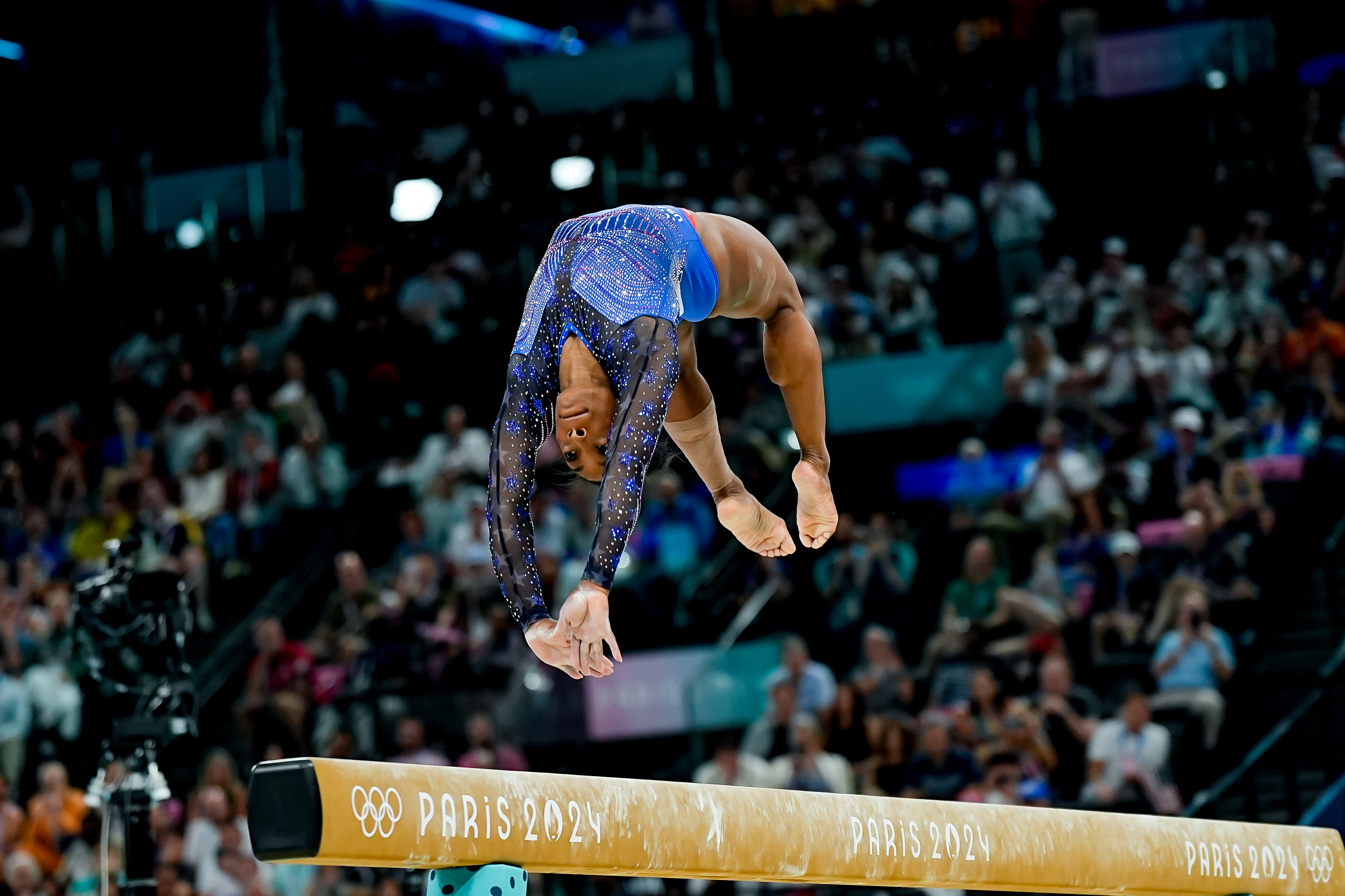 Simone Biles performs a backflip on the balance beam during the Paris 2024 Olympics, surrounded by a cheering crowd
