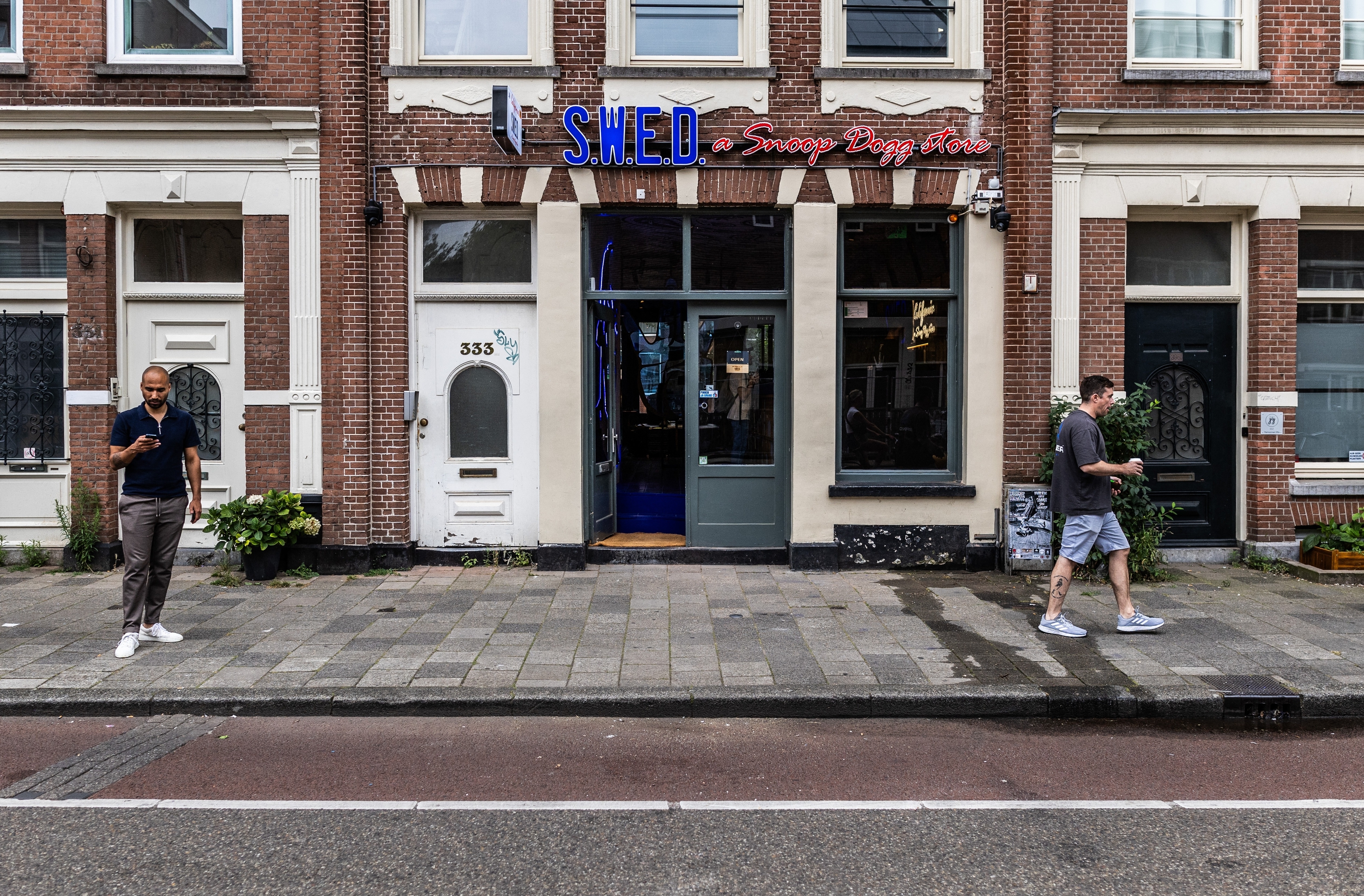Two men stand outside &quot;SHED,&quot; a building with a sign reading, &quot;Snoop Dogg Store,&quot; on a street. Both are looking at their phones