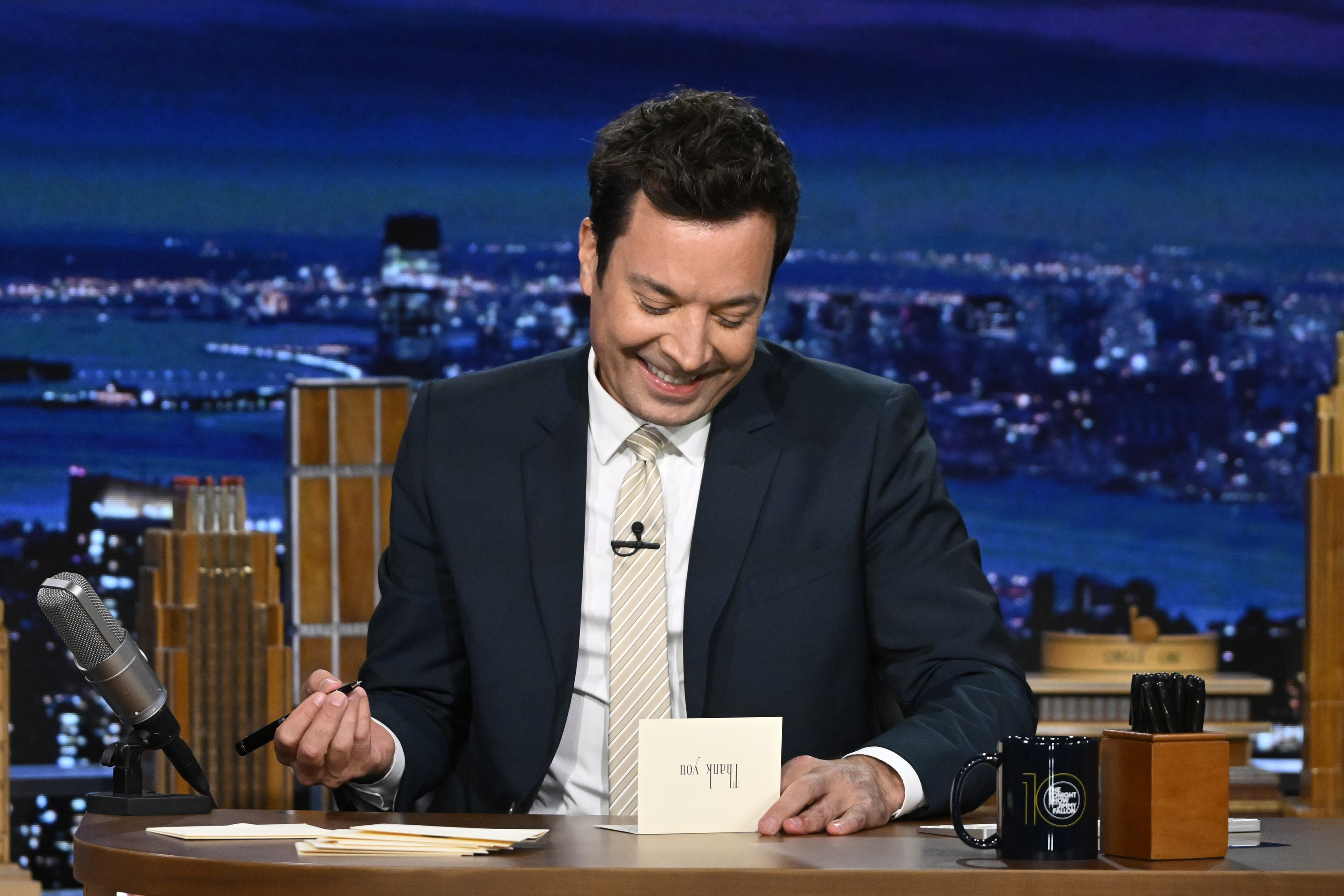 Jimmy Fallon smiles while reading a card on the set of "The Tonight Show" desk, with a cityscape backdrop