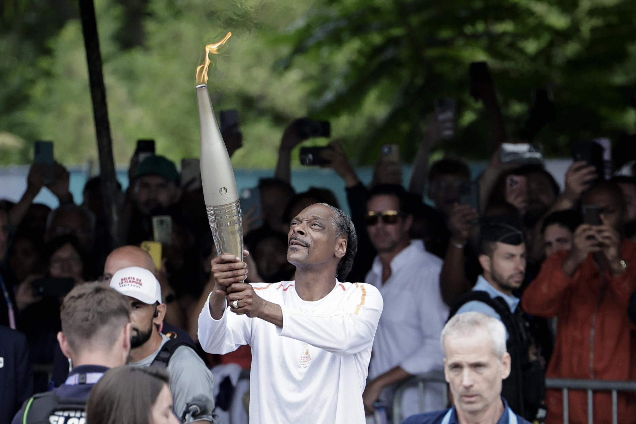 Snoop Dogg, in a white shirt, holds the Olympic torch while surrounded by a crowd taking photos