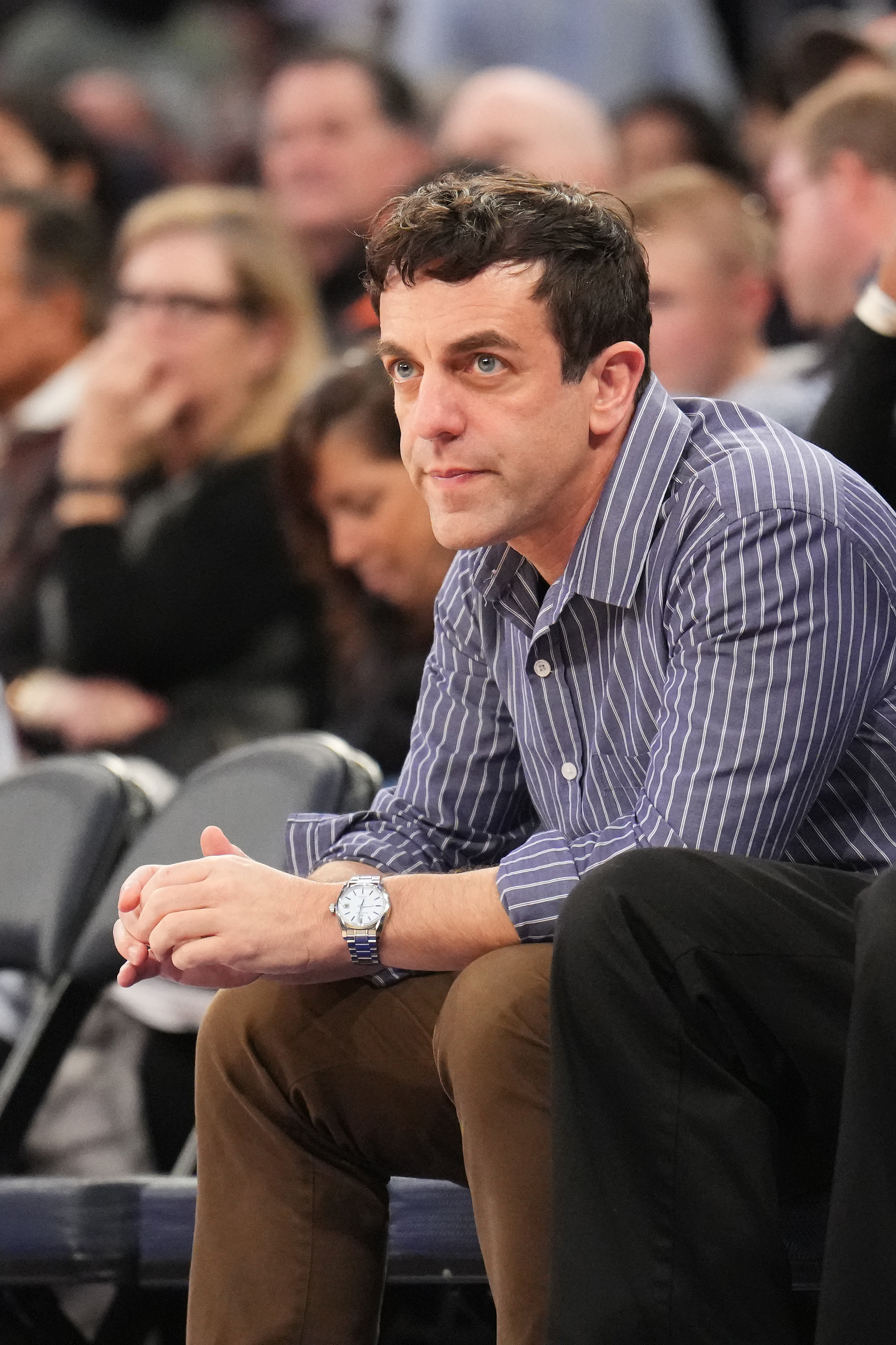 B.J. Novak sits courtside at a basketball game, wearing a striped shirt and brown pants, with a watch on his left wrist, looking focused