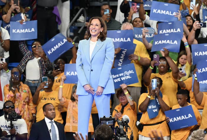 Kamala Harris stands smiling in a light-colored suit at a rally. Supporters hold "KAMALA" signs and cheer in the background
