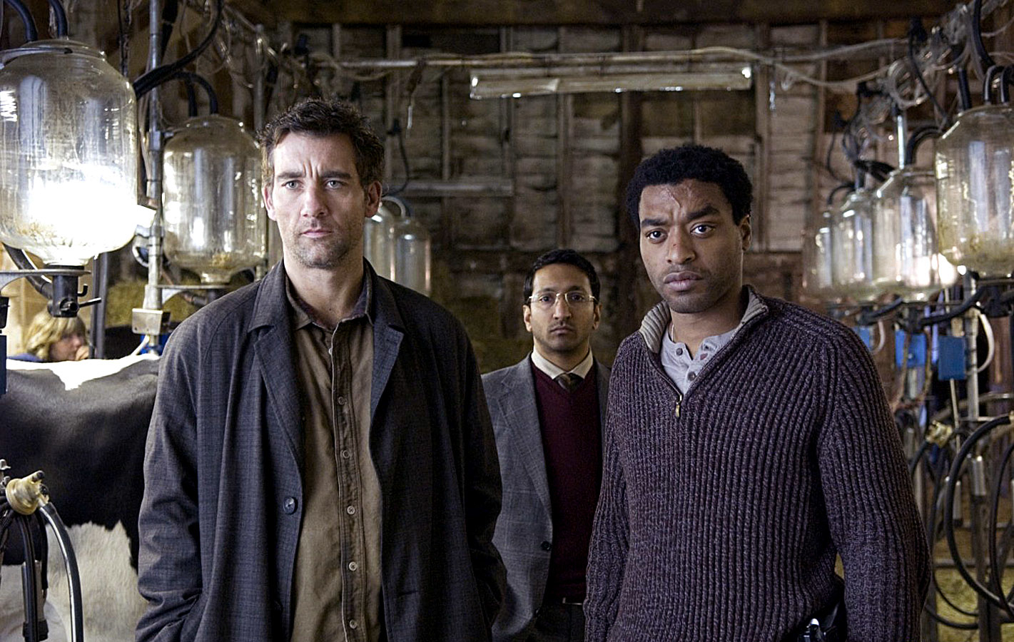 Clive Owen, Chiwetel Ejiofor, and another man stand inside a dimly lit barn with milking equipment in the background