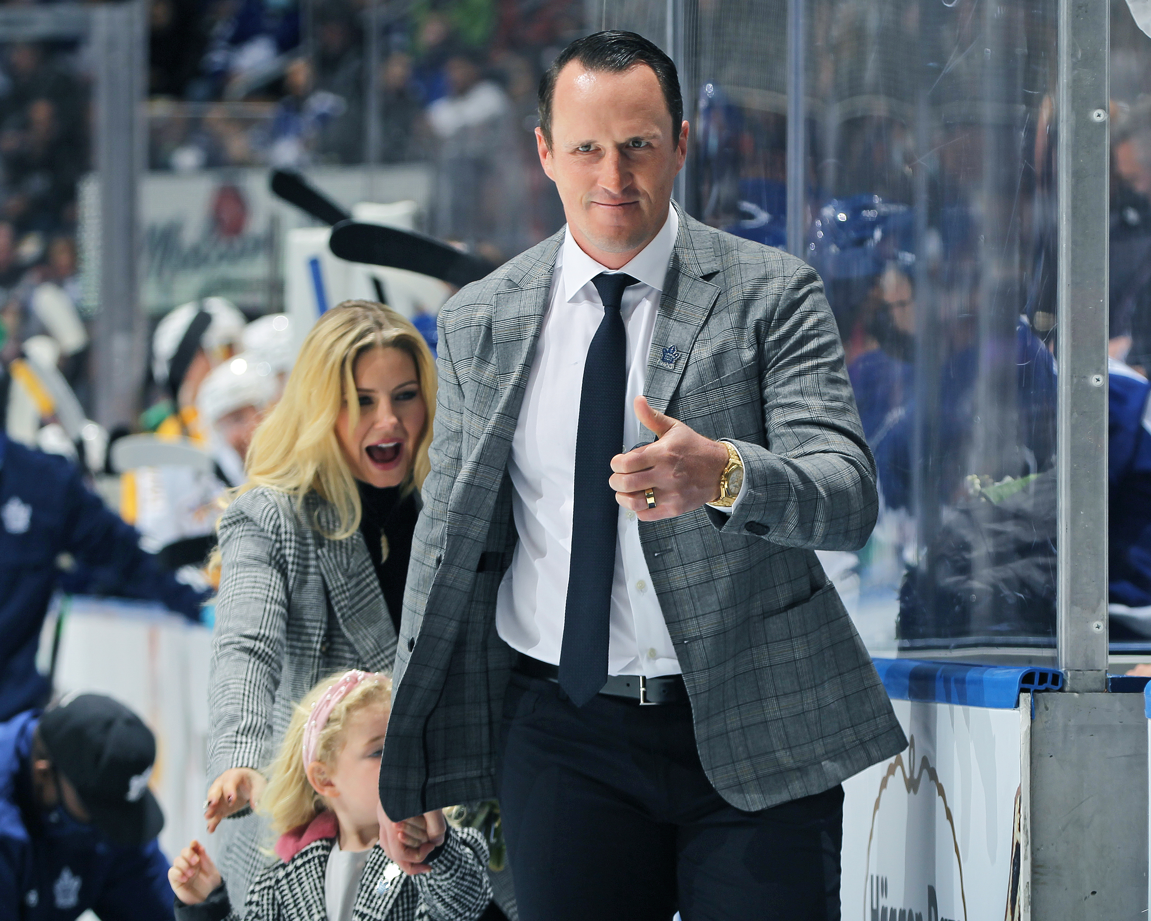 A man in a suit and tie, a woman, and a child stand by a hockey rink. The man gestures with a thumbs-up. The woman appears excited