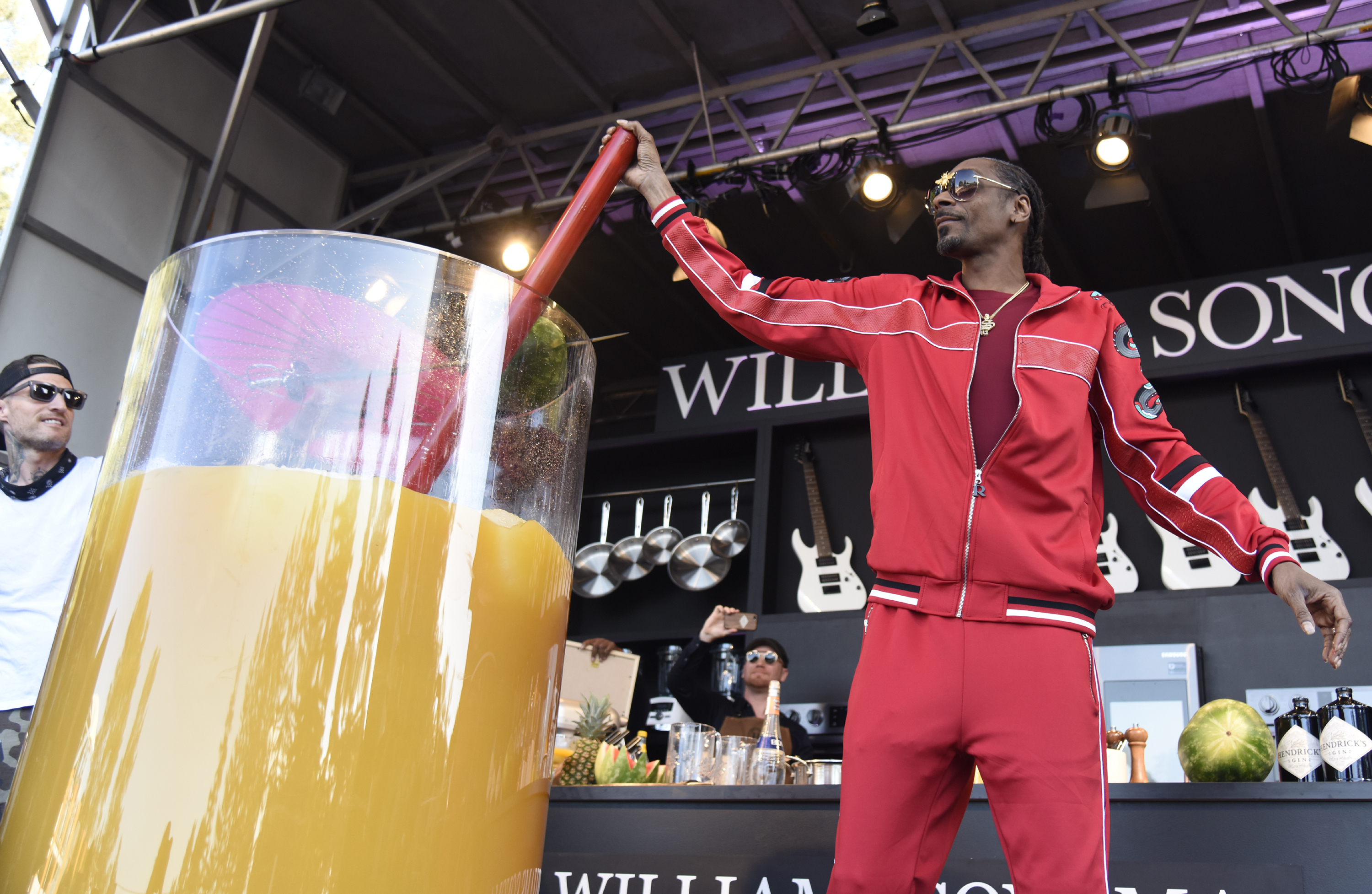 Snoop Dogg, in a red tracksuit, stirs a giant glass of orange drink on stage during a promotional event. Another person looks on from the side