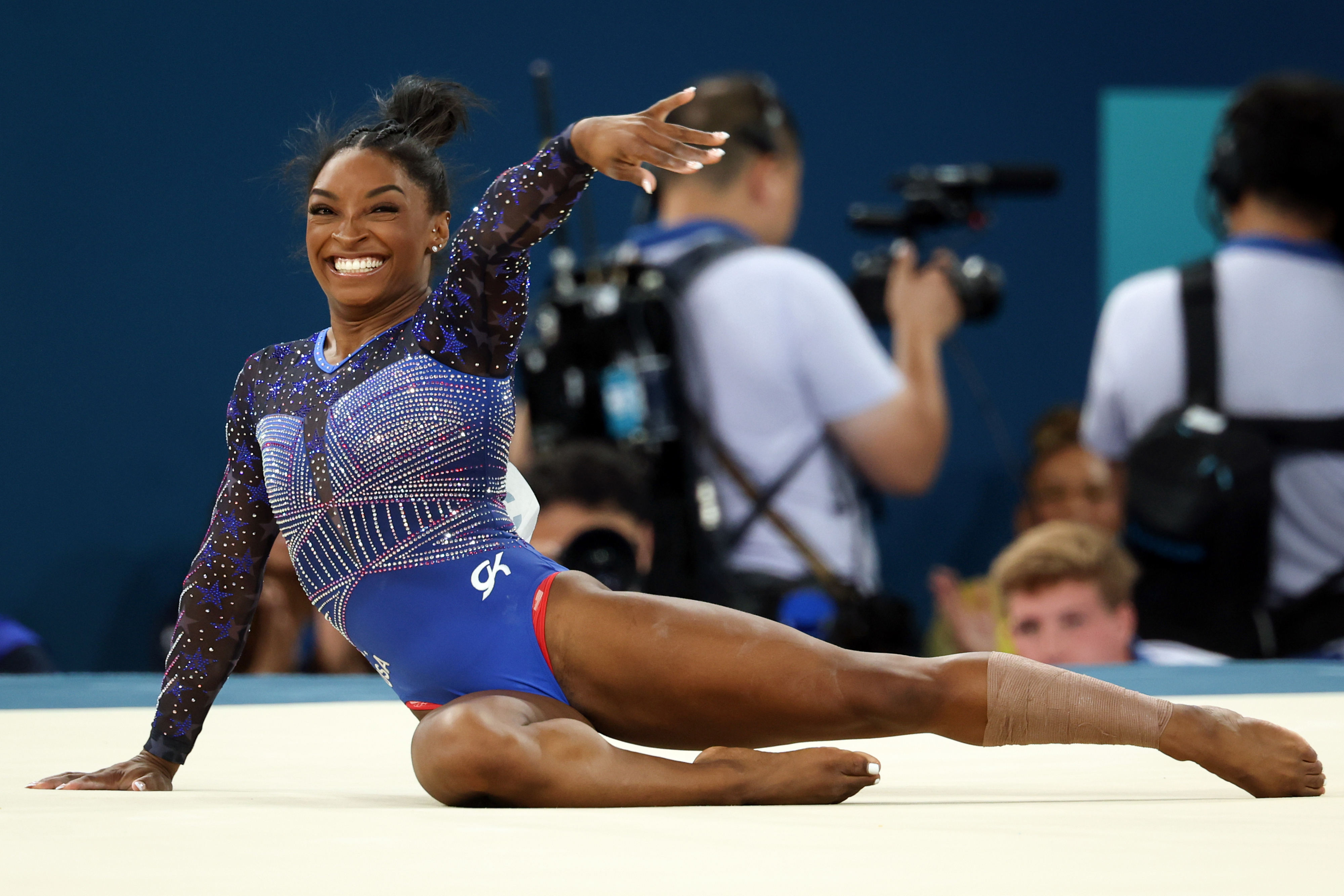 Simone Biles performs a floor routine at a gymnastics competition, wearing a sparkling leotard. She is smiling and posing elegantly with one arm extended